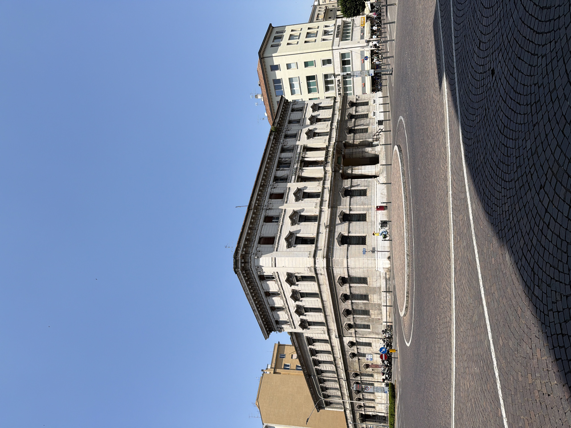 The image shows a street scene in Mantua, Italy. A large, multi-story building with ornate architectural details dominates the view. The building features multiple rows of windows and decorative elements. The street is paved with cobblestones and features painted white lines. Several parked motorcycles line the street near the building. In the background, another building is visible. The sky is clear and blue.
