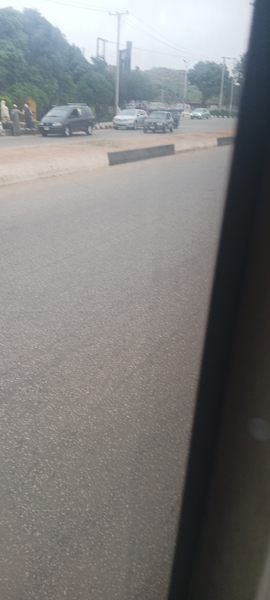 The image captures an outdoor scene from the perspective of a vehicle, looking out onto a road under an overcast sky. The right side of the frame is obscured by a dark pillar or window frame of the vehicle from which the photo was taken.
In the foreground, a gray asphalt road stretches diagonally across the image. A low concrete curb or median runs parallel to the road on the left, separating it from a dirt shoulder or sidewalk where several people are gathered. On the far left, multiple individuals are visible; some are wearing long, light-colored garments (possibly robes or traditional attire), while others are in darker clothing. They appear to be standing or interacting near a dark-colored minivan or SUV, which is either parked or moving slowly on the side of the road. This vehicle has a roof rack.
Further down the road, traffic is present. A light-colored sedan (possibly silver or white) is in the middle lane, followed by a darker sedan. Another light-colored car is visible in the distance. Utility poles with overhead lines stand prominently on both sides of the road in the midground. Dense green vegetation and trees line the left side of the road, and some sparse foliage is visible on the right. A dark, indistinct structure can be seen behind the utility poles in the background.
The overall lighting is diffused, suggesting a cloudy day. No discernable text is visible on any objects, vehicles, or signs. The scene appears to depict a typical street or road in a populated area.