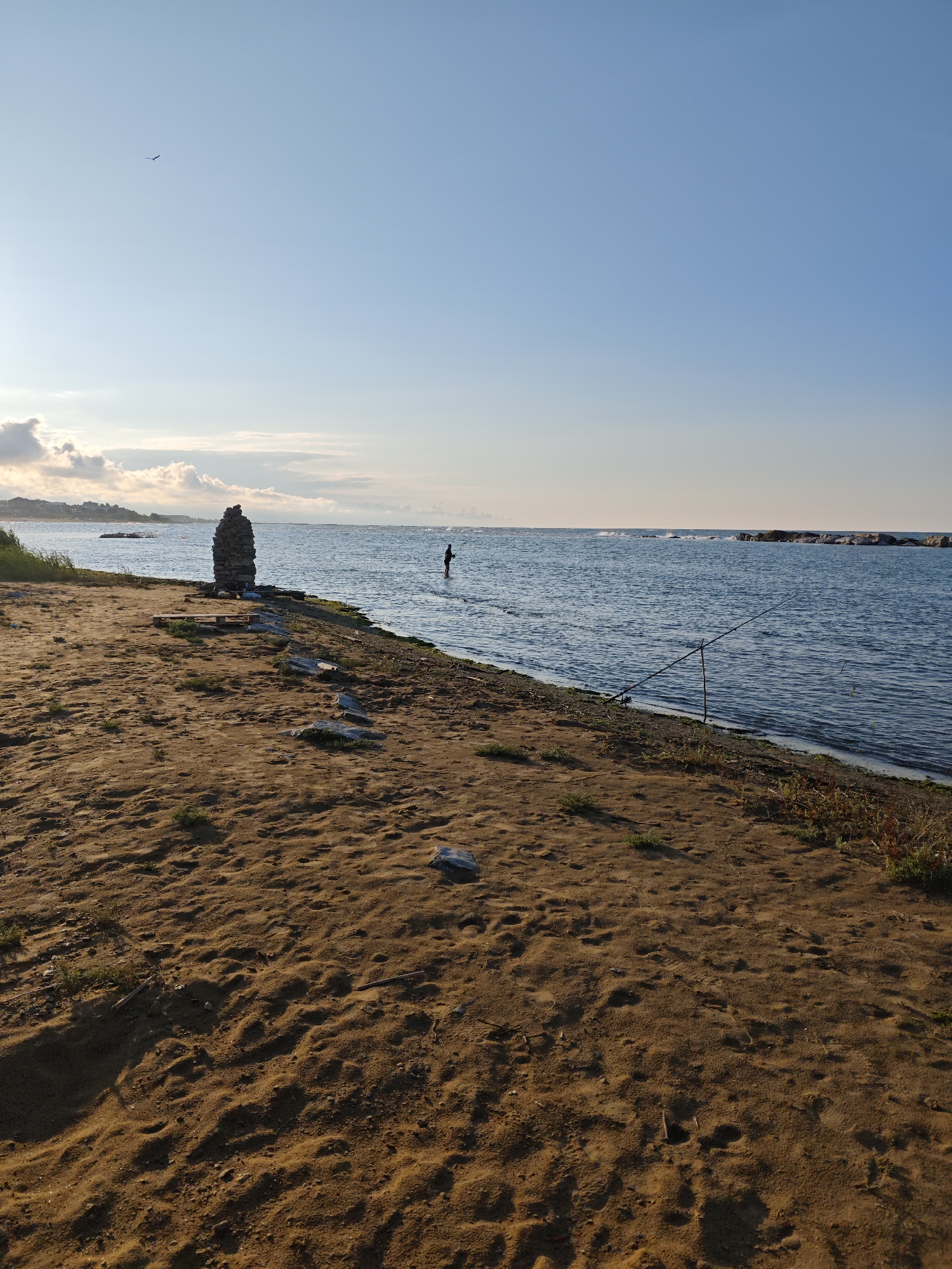 The image depicts a serene coastal scene at Izberbash, Russia, likely during the early morning or late afternoon given the soft, warm lighting. The foreground is dominated by a sandy beach with scattered rocks and vegetation. A tall, stacked stone cairn stands prominently on the shore, serving as a natural landmark.
In the shallow waters of the sea, a lone figure is visible, appearing to be fishing or wading. A fishing rod, propped on a stand, is also present near the water's edge, suggesting fishing activity. The sea itself is calm with gentle waves breaking against a distant breakwater constructed of large rocks.
The sky is a clear, pale blue, with a few wispy clouds on the horizon, hinting at fair weather. A single bird is captured in flight against the expansive sky. In the distance, a town or village with buildings can be seen nestled along the coastline. The overall atmosphere is tranquil and peaceful, evoking a sense of solitude and connection with nature. There is no visible text in the image.