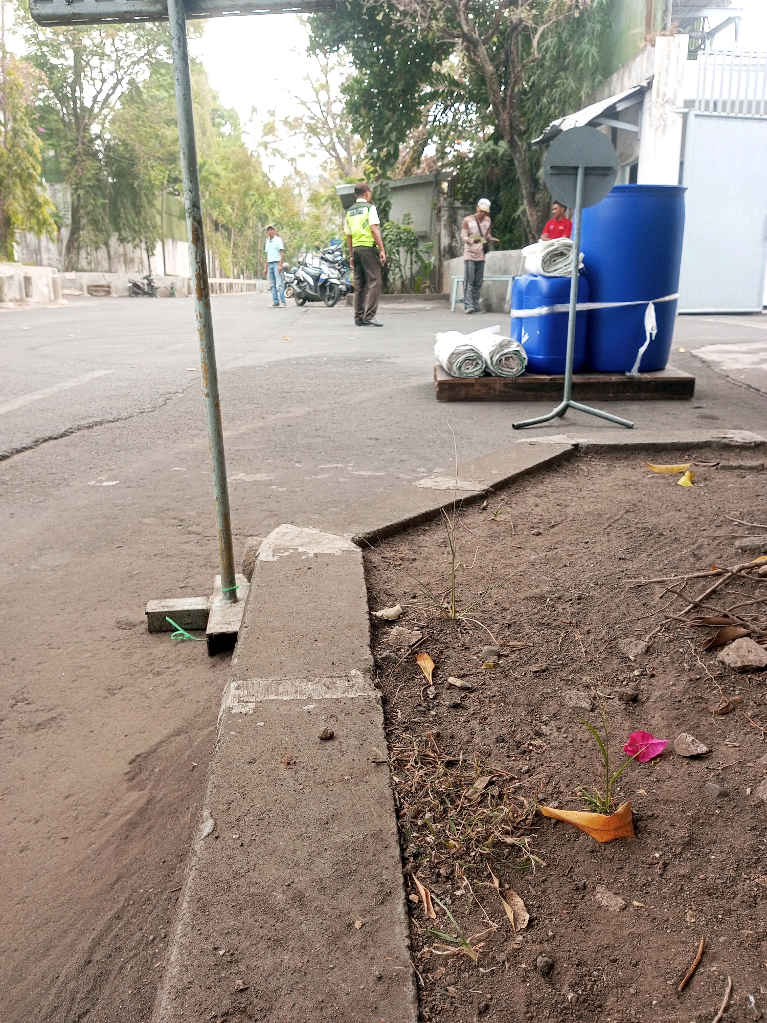 The image captures a street scene in Malang, Indonesia. In the foreground, a concrete curb with dirt and some dried leaves is visible. A thin, weathered metal pole with a green tie is anchored to the ground on the left side of the curb.
Further into the scene, a paved road stretches towards the background. On the road, two large blue plastic barrels are stacked on a wooden pallet, secured with white straps. Rolled-up white fabric or tarps are placed next to the barrels. A metal stand with three legs supports a circular grey sign.
Several people are present. One person in a yellow vest and dark pants stands near the barrels, facing away from the camera. Another person in a light blue shirt and jeans is standing a short distance away, looking towards the barrels. In the background, another individual in a white hat and patterned shirt appears to be holding something. Further back, a person in a red shirt is also visible.
Motorcycles are parked on the side of the road in the middle ground, with trees and buildings forming the backdrop. The overall environment suggests an outdoor, possibly industrial or commercial, area within the city.