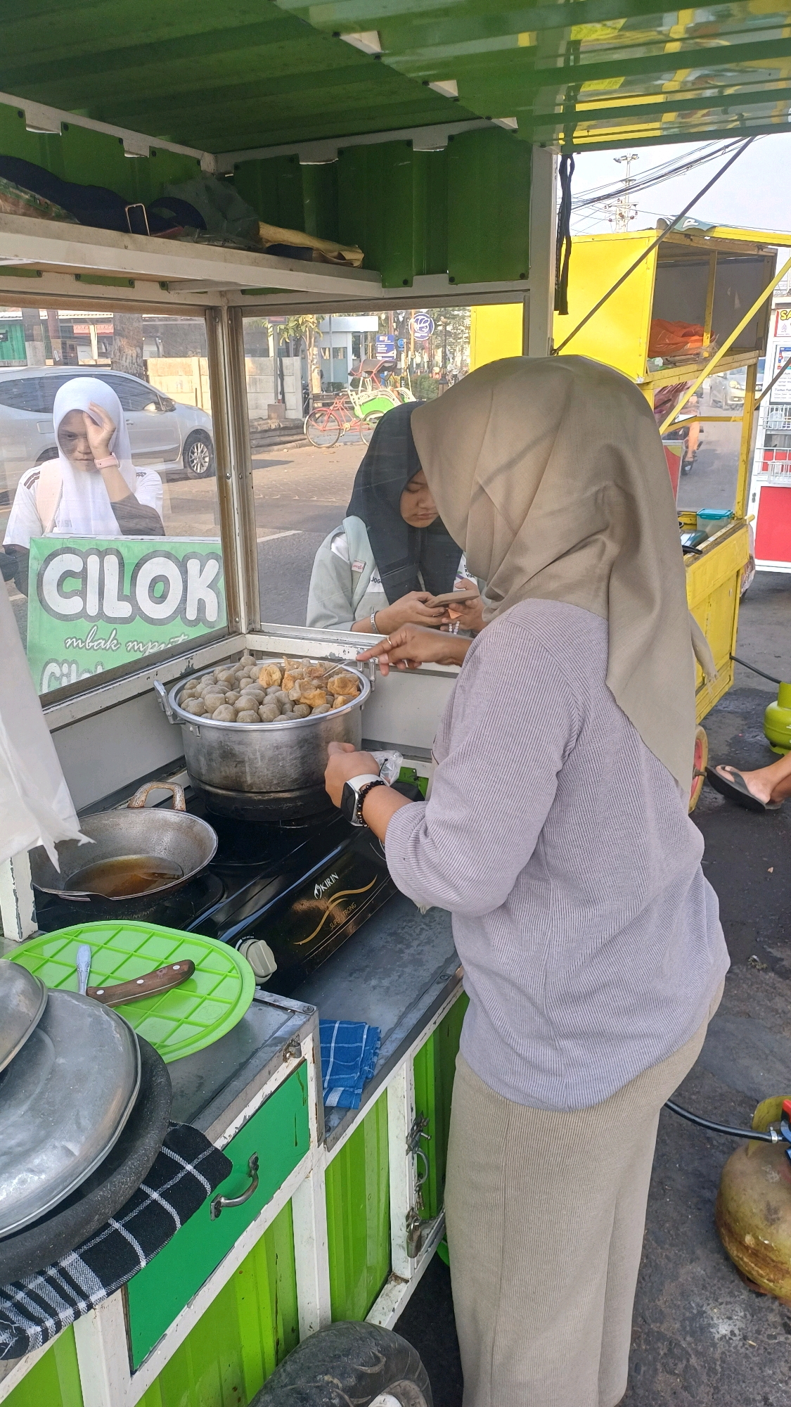 The image depicts a street food stall in Klaten, Indonesia, specializing in "cilok," a type of Indonesian meatballs. A woman wearing a light brown hijab and a striped blouse stands at the counter, seemingly preparing food. In front of her, a large pot filled with cilok sits on a portable gas stove. To her left, a small pan with oil and a green plate with a knife are visible. Behind the counter, two young women in hijabs are visible through the glass. One, wearing a white hijab, looks towards the camera, while the other, in a black hijab, is looking down at her phone. The stall itself is green and white, with a yellow structure behind it, possibly a larger food cart or vehicle. The background shows a street scene with a parked car and a bicycle with a basket. The overall environment suggests a typical urban setting in Indonesia.