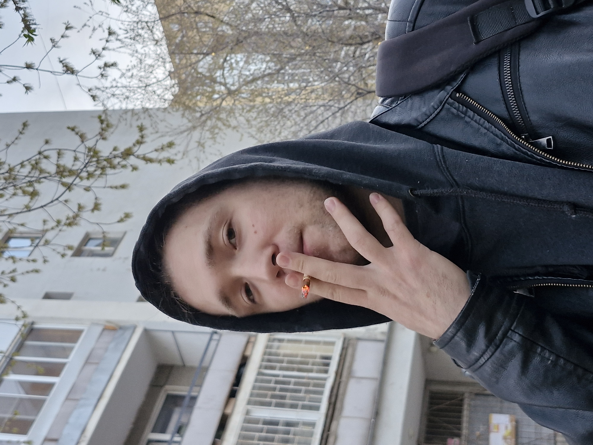 A rotated portrait-oriented image shows a young person in an urban setting, positioned sideways. The subject, appearing to be male, has light skin, short dark hair (partially visible under a hood), and a neutral to slightly contemplative expression, looking directly towards the camera. He is wearing a dark, possibly black, hooded sweatshirt with the hood up, and a black leather jacket with visible zippers. A black backpack strap is seen over his right shoulder.
The person is holding a lit cigarette between the fingers of his left hand, positioned near his mouth. The tip of the cigarette glows orange.
The background features a light-colored, multi-story building with numerous windows, suggesting an apartment block or an institutional building. Some windows are reflective, mirroring tree branches and the sky. Above and behind the subject, bare tree branches with nascent green leaves are visible against an overcast sky. The overall environment suggests an early spring day. The architecture and general atmosphere are consistent with an urban area in Yekaterinburg, Russia. There is no visible text.