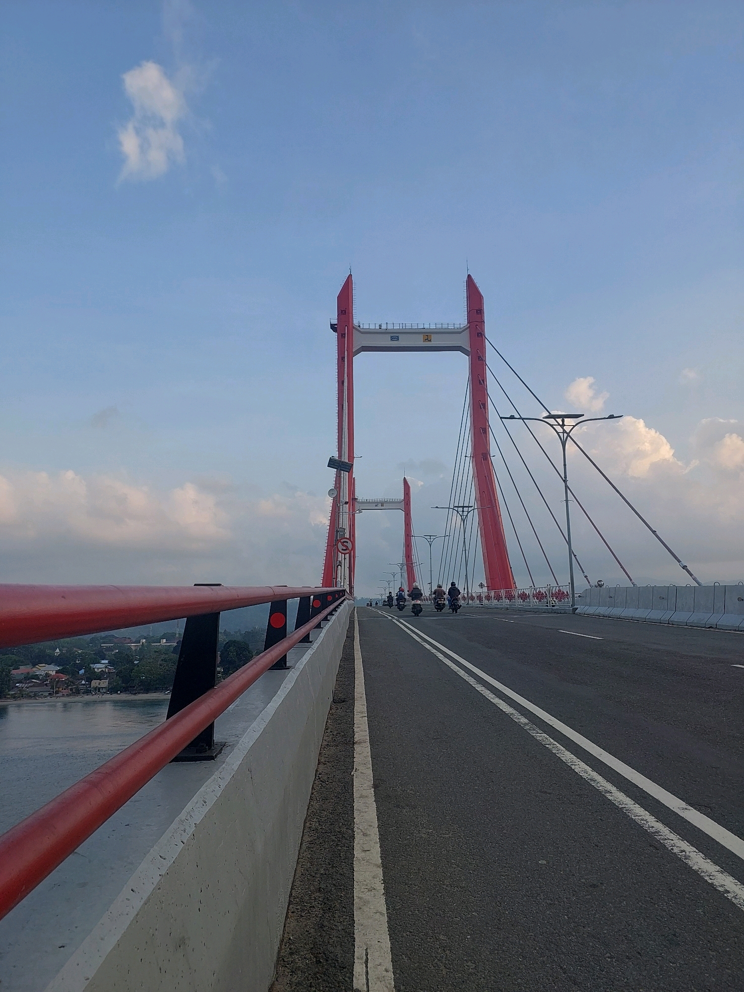 The image depicts a view from a cable-stayed bridge, likely the Youtefa Bridge in Ambon, Indonesia, during daylight hours under a partly cloudy sky. The bridge has distinctive red towers and cables, with a paved road surface marked with white lines. A red railing runs along the left side of the bridge, overlooking a body of water and a densely populated area with houses and trees. Several motorcycles carrying riders are traveling on the bridge in the distance, indicating ongoing traffic. The overall scene suggests a functional infrastructure element integrated into the urban landscape of Ambon.