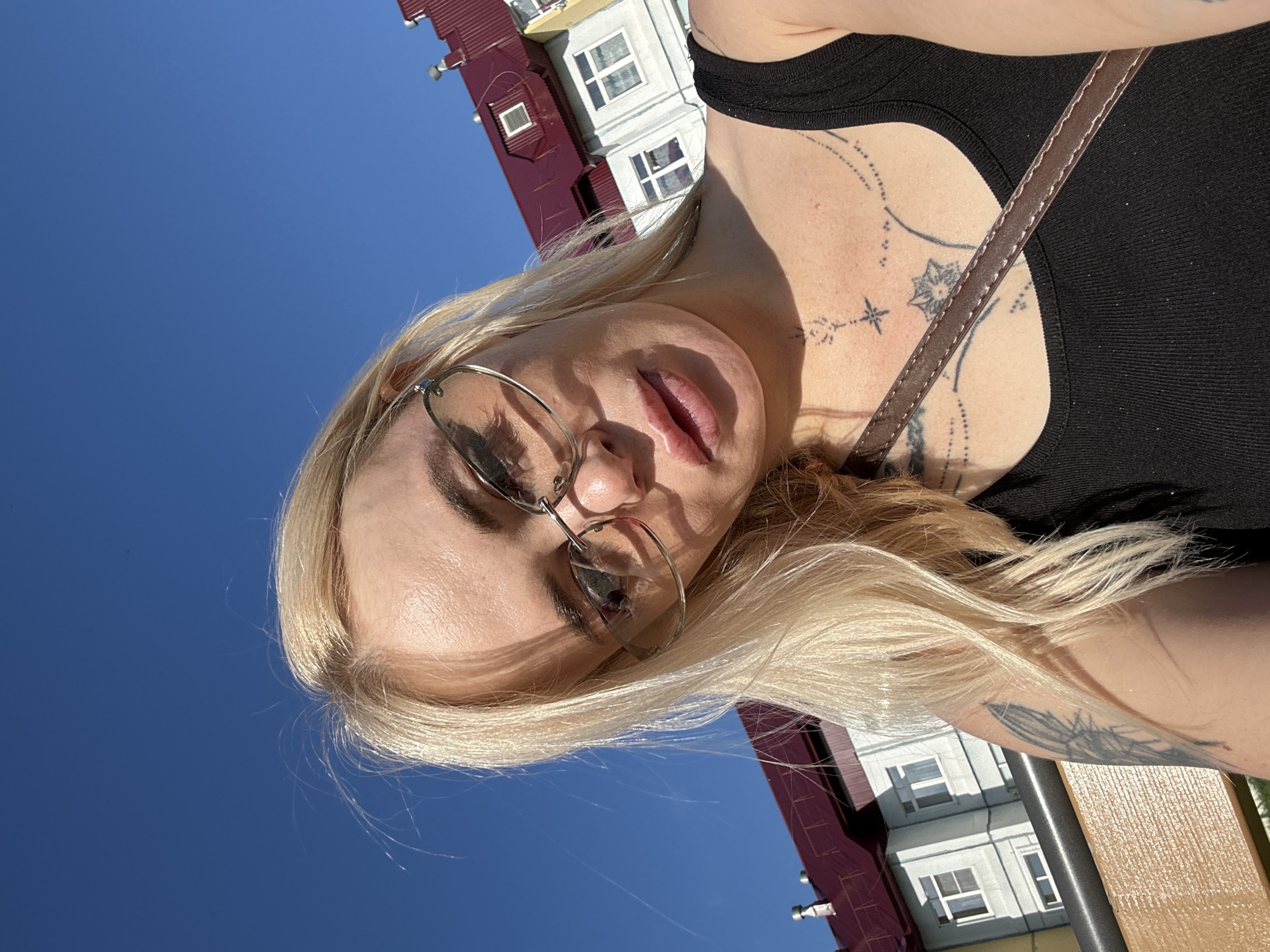 A woman with blonde hair and glasses is taking a selfie outdoors. She is wearing a black tank top and has tattoos on her chest and arm. The background shows a bright blue sky and the exterior of a building with a red roof. The woman appears to be sitting on a bench.