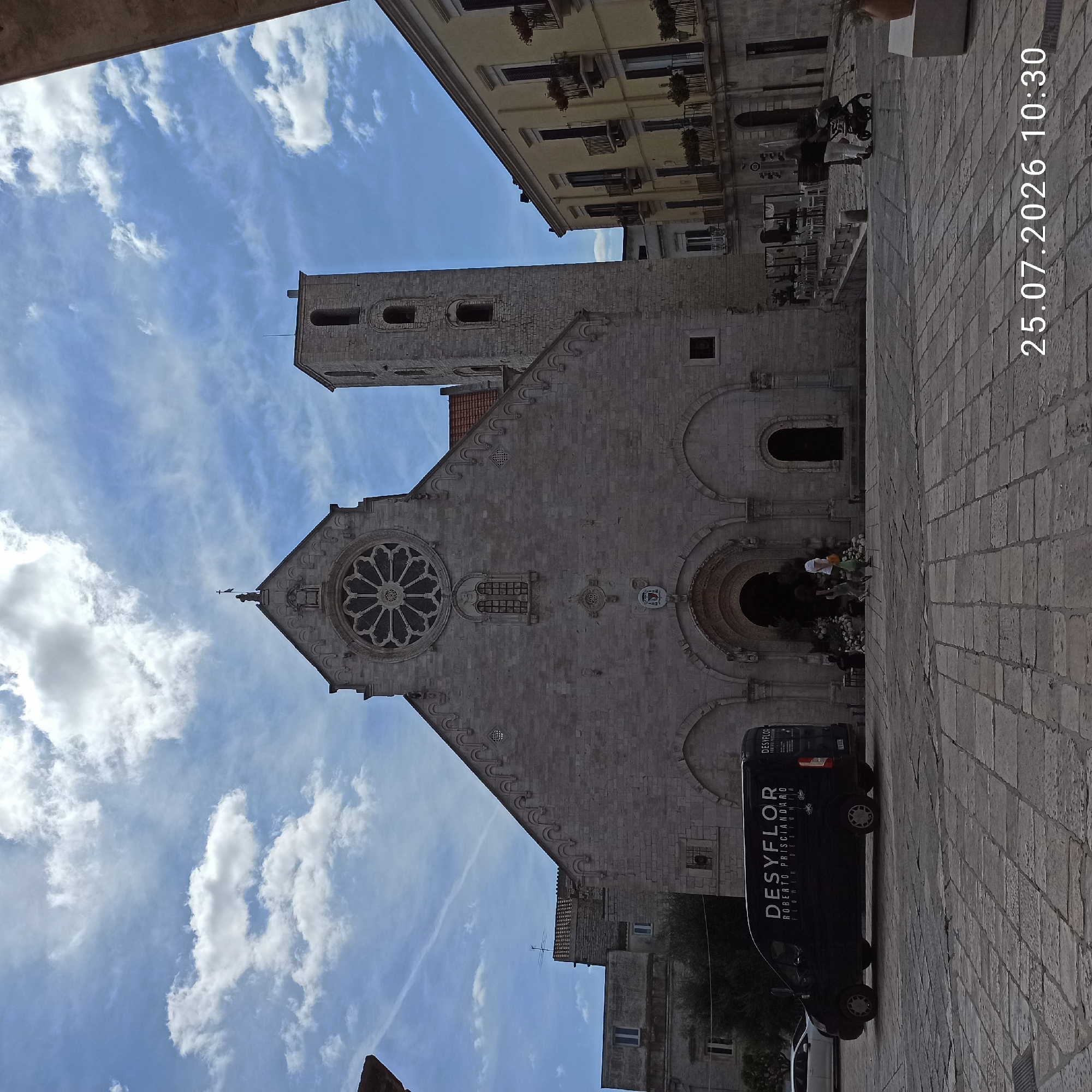 This is a wide-angle photograph taken on a sunny day in front of a large stone church, identified as the Duomo di Ruvo di Puglia, Italy. The church features a prominent rose window on its facade and a tall bell tower to its right. In the foreground, a black commercial van with the branding "DESYFLOR ROBERTO PRISCIANDARRO FLOWER DESIGNER" is parked on a cobblestone plaza. Two people are walking towards the church entrance, and further to the right, another person is pushing a stroller. The sky is blue with scattered white clouds, and adjacent buildings, including a yellow one with balconies, line the right side of the plaza. A date and time stamp, "25.07.2026 10:30", is visible in the bottom right corner.