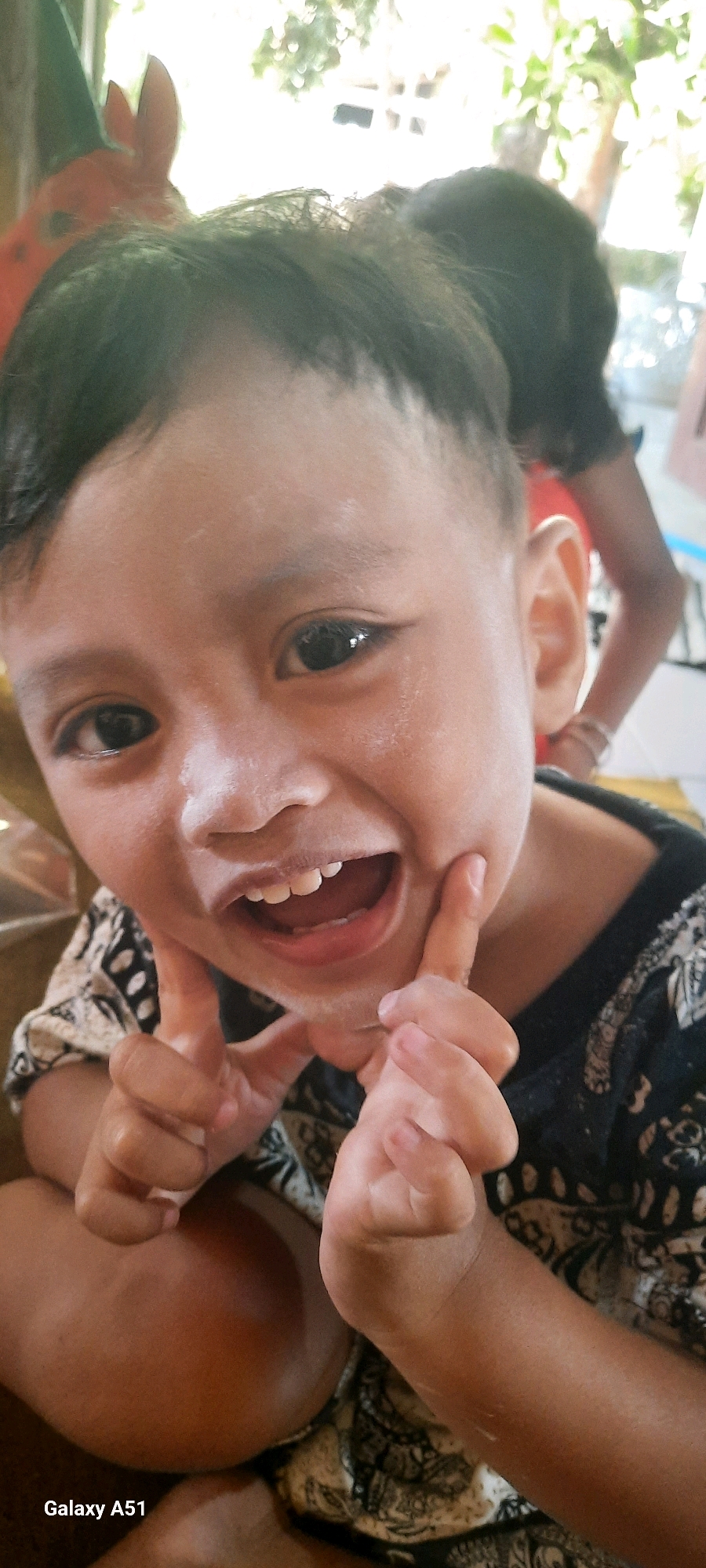 A close-up, frontal, eye-level shot captures a young child with dark hair, light skin, and wide, dark eyes, smiling with their mouth open, showing their teeth. The child is wearing a dark shirt with a white patterned design and has their fingers placed on their cheeks. The background is blurred, showing greenery and a glimpse of another person. The bottom left corner of the image displays "Galaxy A51", indicating the device used for capture. The overall scene suggests a casual, indoor or semi-outdoor setting, potentially in a residential area of Ploso, Indonesia, given the context of the original request.