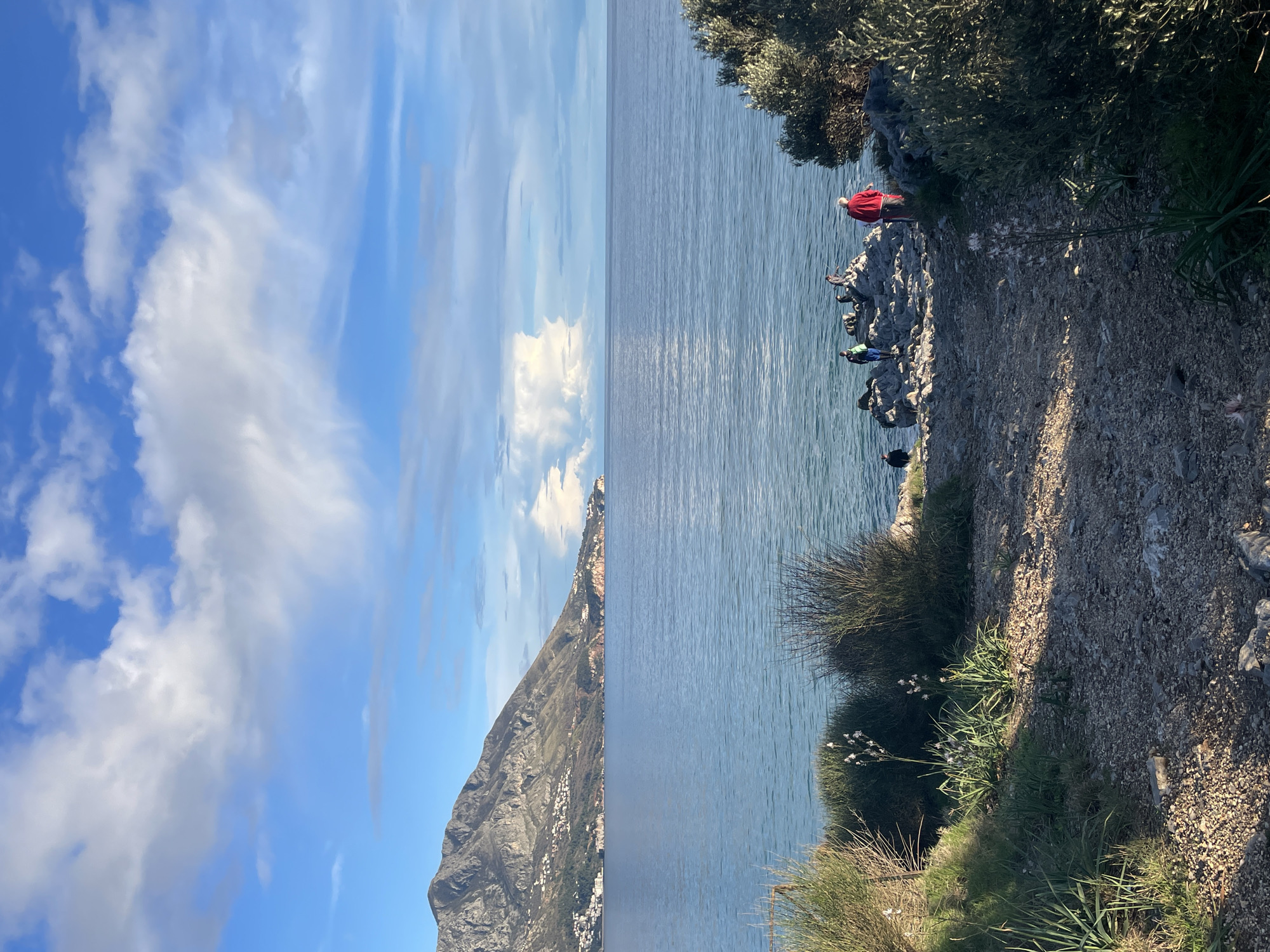The image displays a coastal landscape in Boreti, Montenegro, under a partially cloudy blue sky. The Adriatic Sea occupies the middle ground, its surface calm. To the left, a large, rugged mountain range slopes towards the sea, featuring numerous white buildings scattered across its lower reaches. The foreground comprises a downward-sloping, unpaved path made of small rocks and dirt, flanked by green grass and dense, dark green Mediterranean scrub. Several tall plants with white flowers are visible amidst the vegetation.
Five individuals are present on the rocky shoreline where the path meets the water. On the right, one person in a red top stands on a dark rock, facing the sea. To their left, a group of approximately three people are positioned on dark rocks near the water, one wearing a blue top and green lower garment. A fifth person, dressed in dark clothing, is partially obscured further left near the water's edge. Their actions are consistent with recreational activity along the coast. Long shadows suggest the sun is low in the sky.