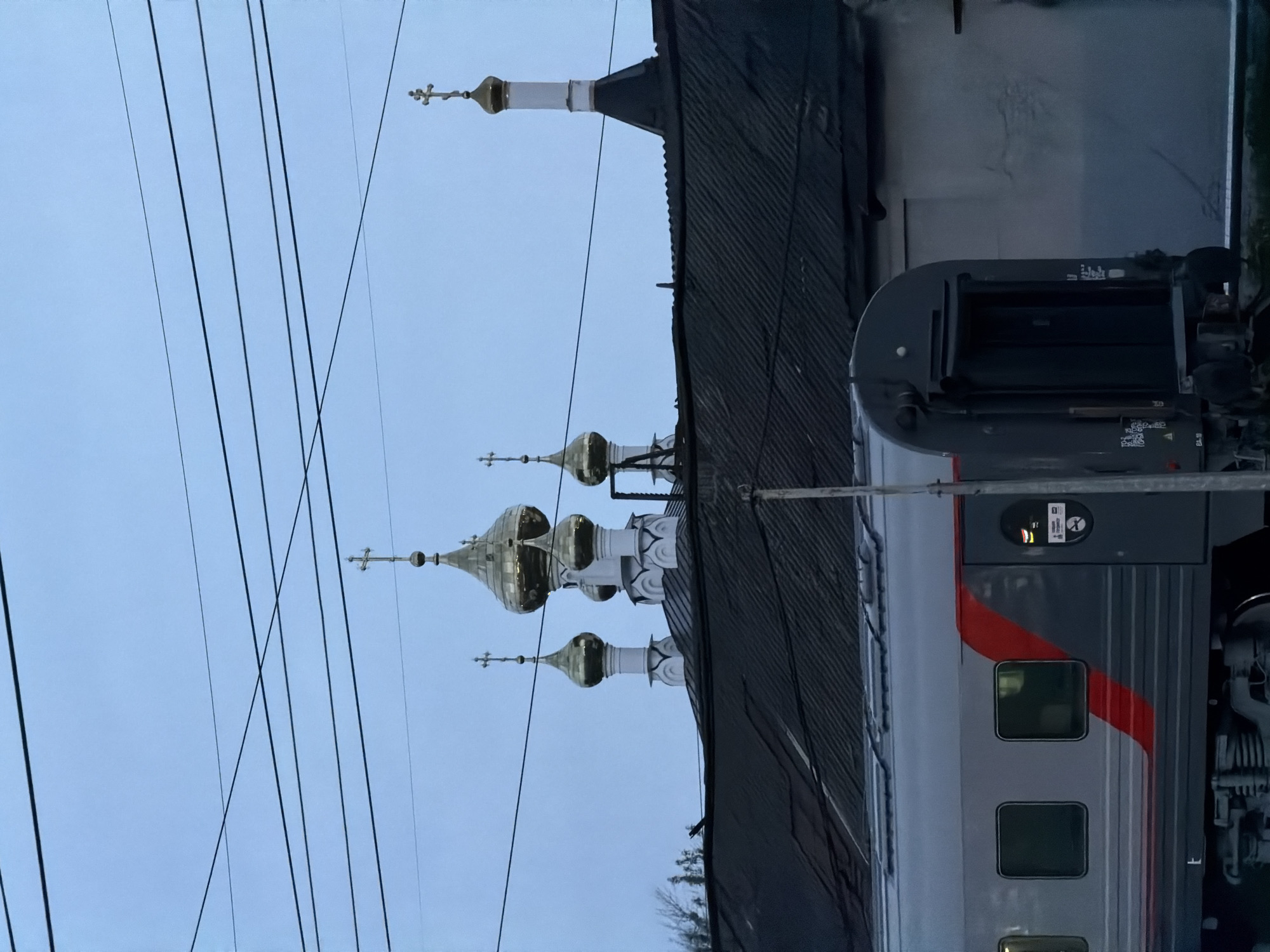 The image depicts a scene in Rtishchevo, Russia, juxtaposing a railway environment with the distinctive architecture of Russian Orthodox churches. In the foreground, a passenger train car is visible, marked with a red stripe, indicating that this is a train in Russia. In the background, the golden domes of a church are visible, towering above the train. Power lines intersect the view, spanning the sky between the train and the church domes. The photograph is taken in the evening, given the low light and the dark clouds.