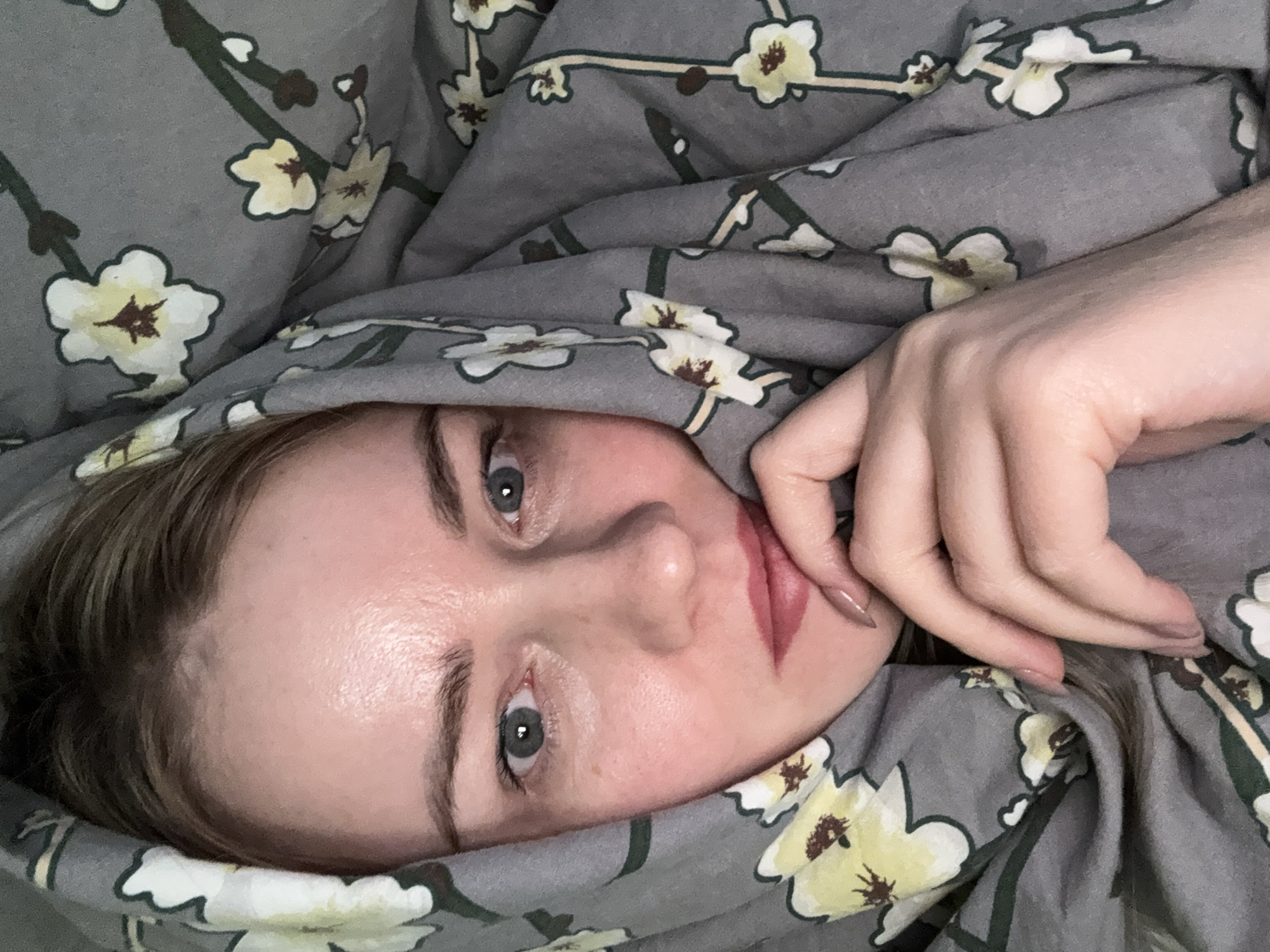 A woman's face is framed by a gray floral patterned bedspread. The woman has fair skin, blue eyes, and light brown hair. Her hand is positioned near her mouth, her index finger touching her lower lip. The background consists of the floral fabric which covers the bedding. This could be a residential setting, possibly a home in Chaikovsky, Russia, as the user specified the location in the request.