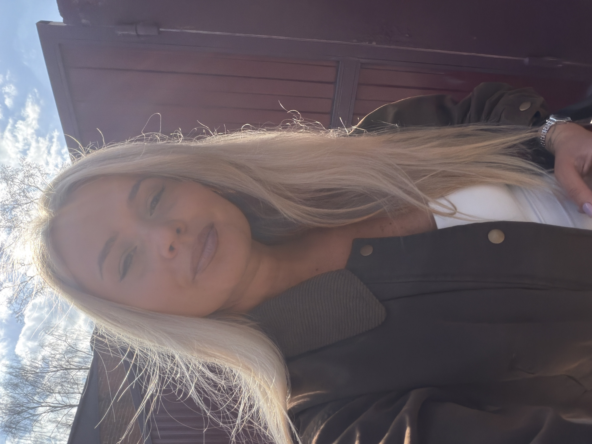 A portrait-style image, likely a selfie, features a young woman with long, straight blonde hair, positioned horizontally. She is wearing a dark olive green or brown bomber-style jacket with snap buttons over a white top. Her face, partially visible and tilted, shows a neutral expression with subtle makeup. Her left hand, visible on the right side of the frame, has light lavender nail polish and a silver-toned watch with a shimmering band.
The background consists of a dark reddish-brown building or wall, possibly corrugated metal or wood siding, below a bright blue sky with scattered white clouds. Bare tree branches are visible in the upper left portion of the image. The scene is outdoors during the daytime under bright, sunny weather. The strong sunlight, likely coming from behind the subject, creates a luminous halo effect on her blonde hair, highlighting individual strands. The overall atmosphere is bright and clear, capturing a casual outdoor moment. No visible text is present.