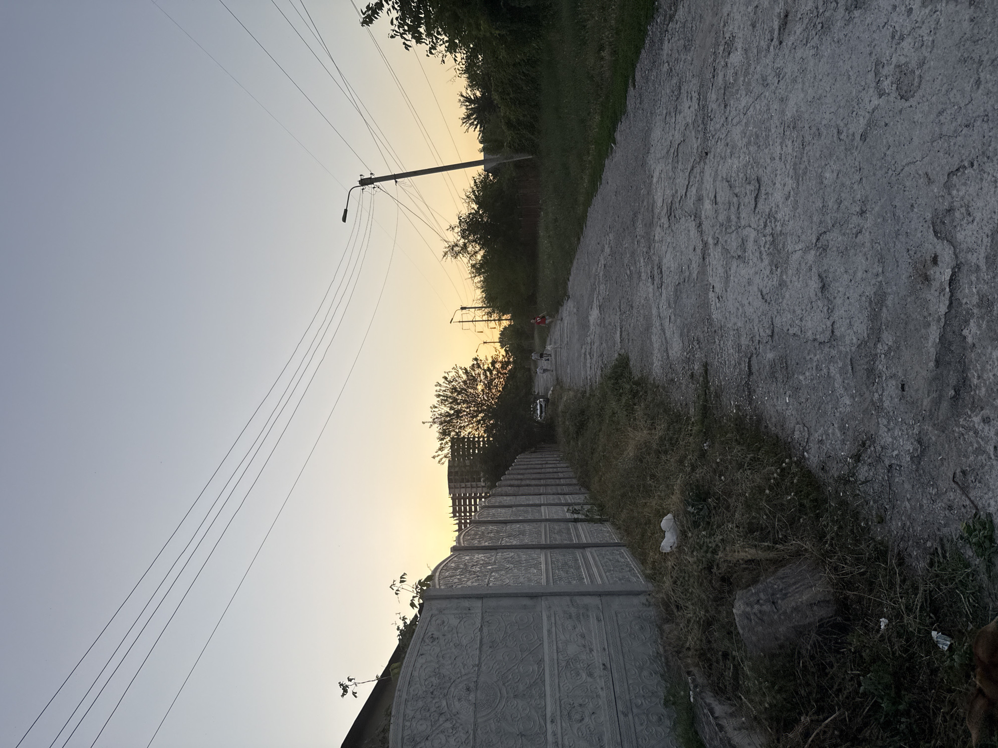 In Zaporizhzhia, Ukraine, at dusk, a dirt road stretches into the distance, flanked by a textured concrete fence on the left and overgrown grass and trees on the right. Power lines crisscross the pale orange and blue sky. A street lamp stands on a pole to the right. In the background, a building under construction is visible. Several people and a dog are walking away from the camera on the road. The area appears to be an unpaved thoroughfare in a semi-developed or rural part of the city.