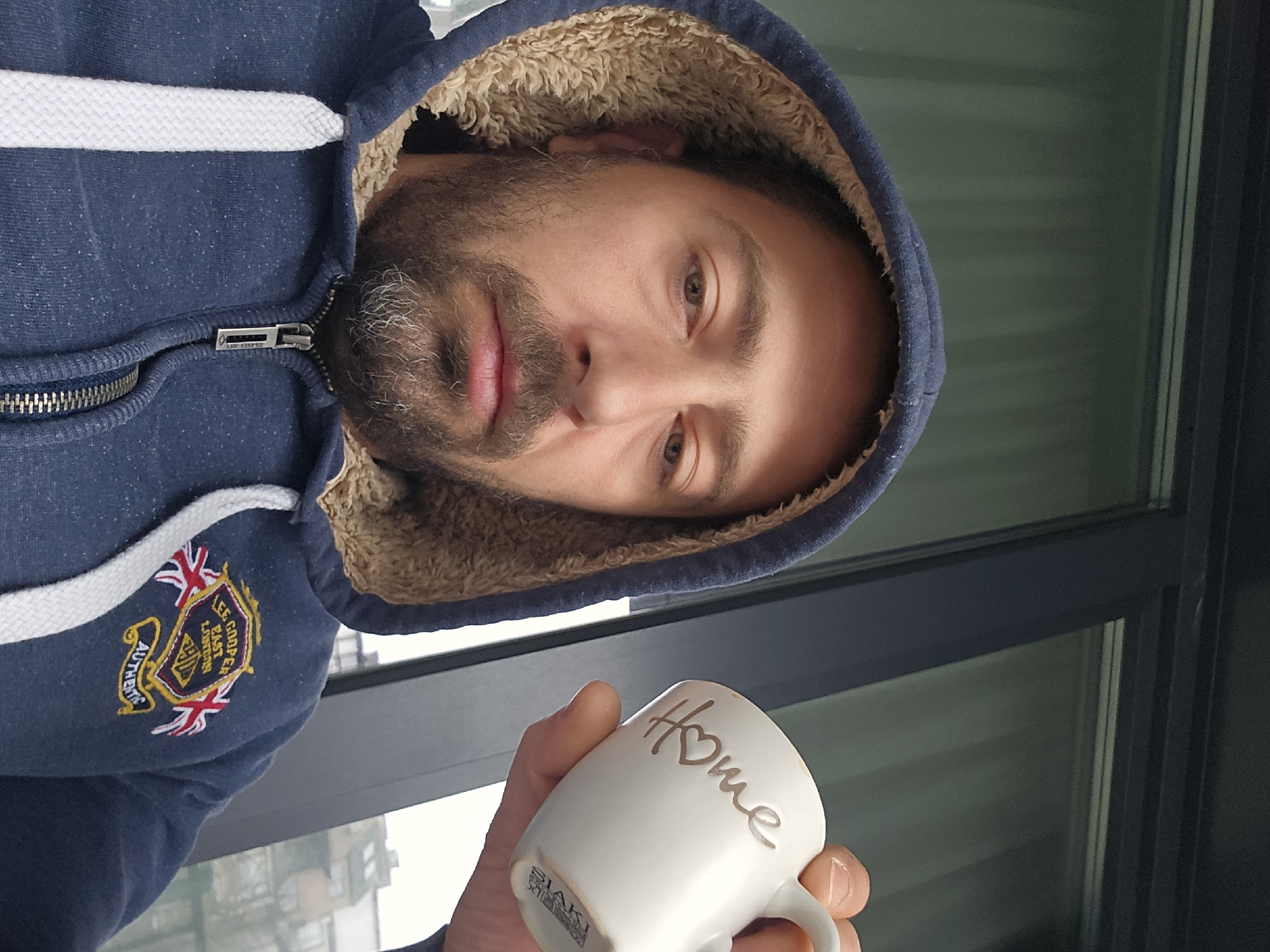 A man with a beard, wearing a blue hoodie with a fur-lined hood, is holding a white mug with the word "Home" inscribed on it. The zipper of the hoodie is visible. The background includes a window, and judging from the exterior view through the window, it appears to be a residential area. It's not possible to verify the location in the city of Burgas, Bulgaria, with the information provided.