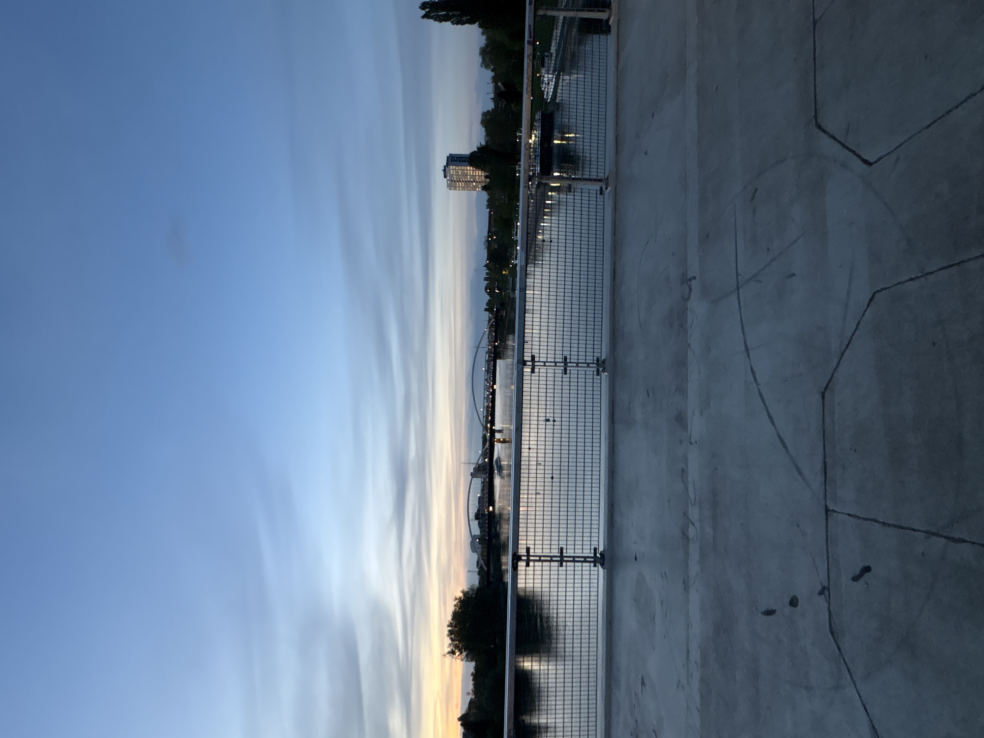 This media file captures a serene urban waterfront scene at twilight or early evening. The image is taken from a raised perspective overlooking a wide body of water, likely a river or canal.
In the foreground, to the right, there is a broad concrete or paved platform with distinct lines and markings. A sturdy metal grid fence runs along the edge of this platform, separating it from the water. The fence appears well-maintained, suggesting a public promenade or viewing area.
Across the water, the far bank features a diverse skyline of buildings. A prominent multi-story building, possibly an apartment complex or office building, stands out with numerous illuminated windows, indicating the onset of night. Other smaller structures and dense trees line the water's edge. In the distance, several arched structures are visible, appearing to be bridges connecting different parts of the city. Reflective lights from the buildings and possibly streetlights create shimmering patterns on the water's surface.
The sky occupies the upper portion of the frame, displaying a gradient from a clear, deep blue at the very top to lighter, warmer tones of pale blue and faint yellow closer to the horizon. Long, wispy, horizontal cloud streaks stretch across the sky, adding texture and depth to the evening light.
There are no people or specific activities discernible. The scene conveys a calm and peaceful atmosphere. No visible text is present in the image. The time of day is clearly evening, with the natural light fading and artificial lights beginning to glow, characteristic of a dusk or early night setting. Given the context of Strasbourg, France, this view likely depicts a part of the city along the Ill River or a canal, with its characteristic bridges and urban architecture.