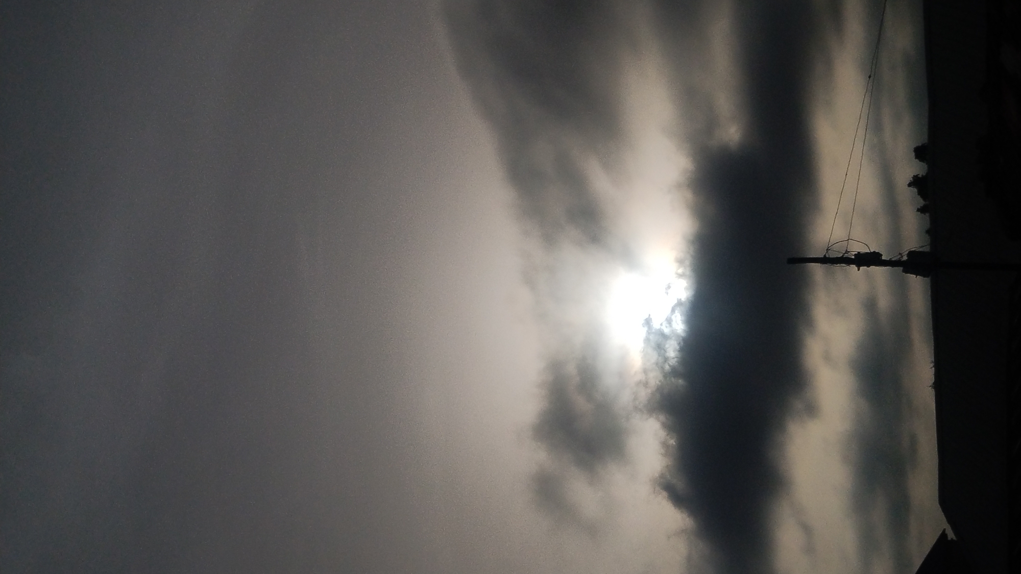 The image shows a daytime scene with a cloudy sky, possibly in Jos, Nigeria. The sun is obscured by thick clouds, creating a diffused light. A utility pole with wires extends vertically on the right side of the frame. Buildings or trees are silhouetted against the sky on the extreme right. The overall impression is overcast and potentially stormy.