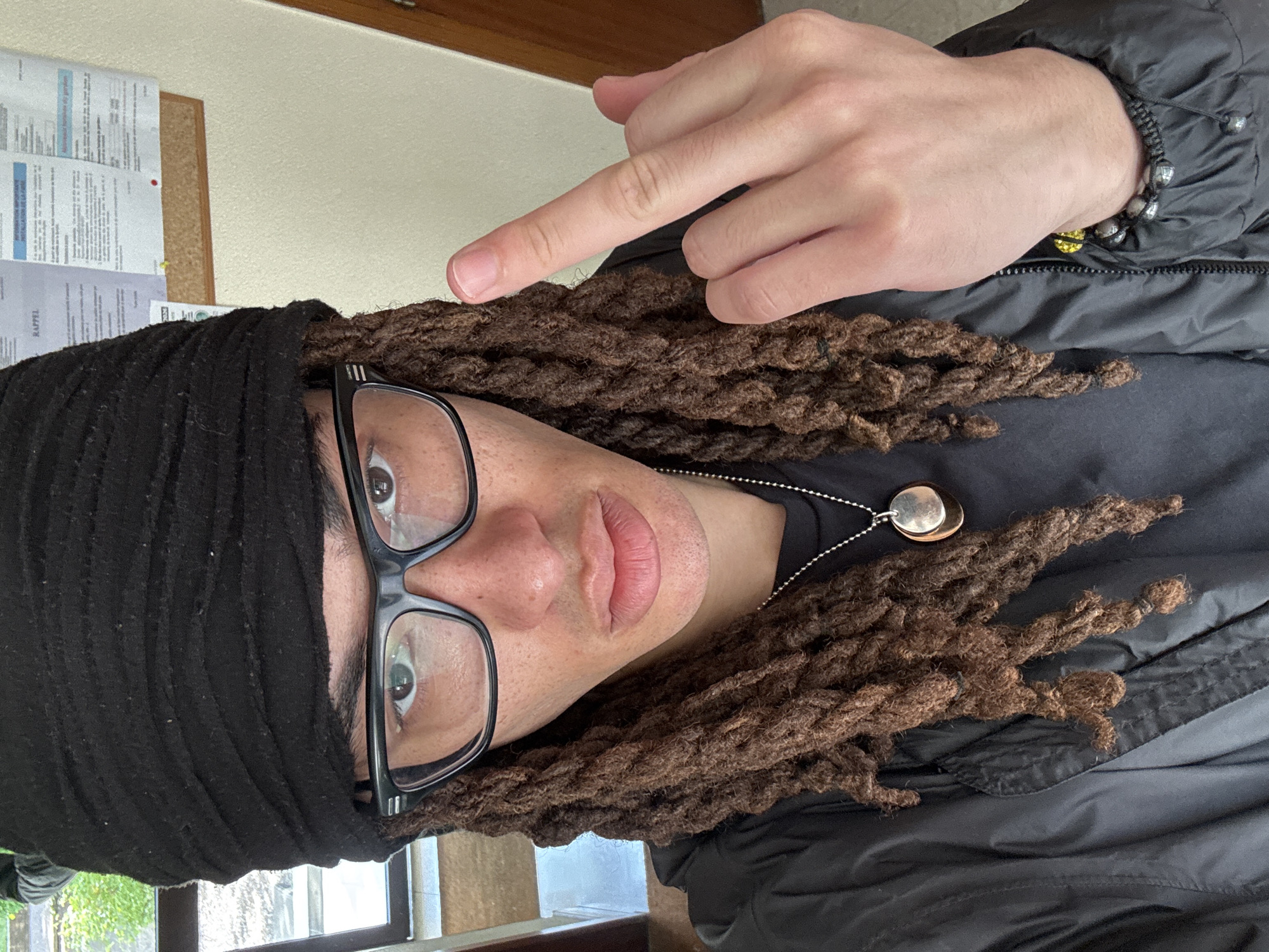 An upright, close-up, eye-level shot captures a person, likely in an indoor setting, making a defiant gesture. The subject's head is tilted slightly, and they are looking directly towards the viewer. Their right hand is raised, extending the middle finger.
The person has brown dreadlocks, which are partially covered by a black head wrap or beanie. They wear black-rimmed glasses, and their light-colored eyes are clearly visible. Their lips are full, and there's some subtle stubble visible on their chin and upper lip area. Around their neck, a silver chain holds a distinctive, circular metallic pendant with an irregular, slightly polished surface. They are dressed in a black, possibly padded or puffer jacket, visible on their right shoulder and arm. The wrist of the gesturing hand features a beaded bracelet, composed of dark, metallic-looking beads strung on a black cord.
In the background, behind the person's left shoulder, is a light-colored wall. A cork board is affixed to this wall, displaying several documents or notices. One of these papers clearly shows the French word "RAPPEL" (meaning "reminder") in capitalized letters. To the bottom left, a window or glass partition offers a blurred view of some outdoor greenery. The overall lighting suggests it is daytime. The gesture, combined with the neutral facial expression, conveys a direct and possibly challenging message.