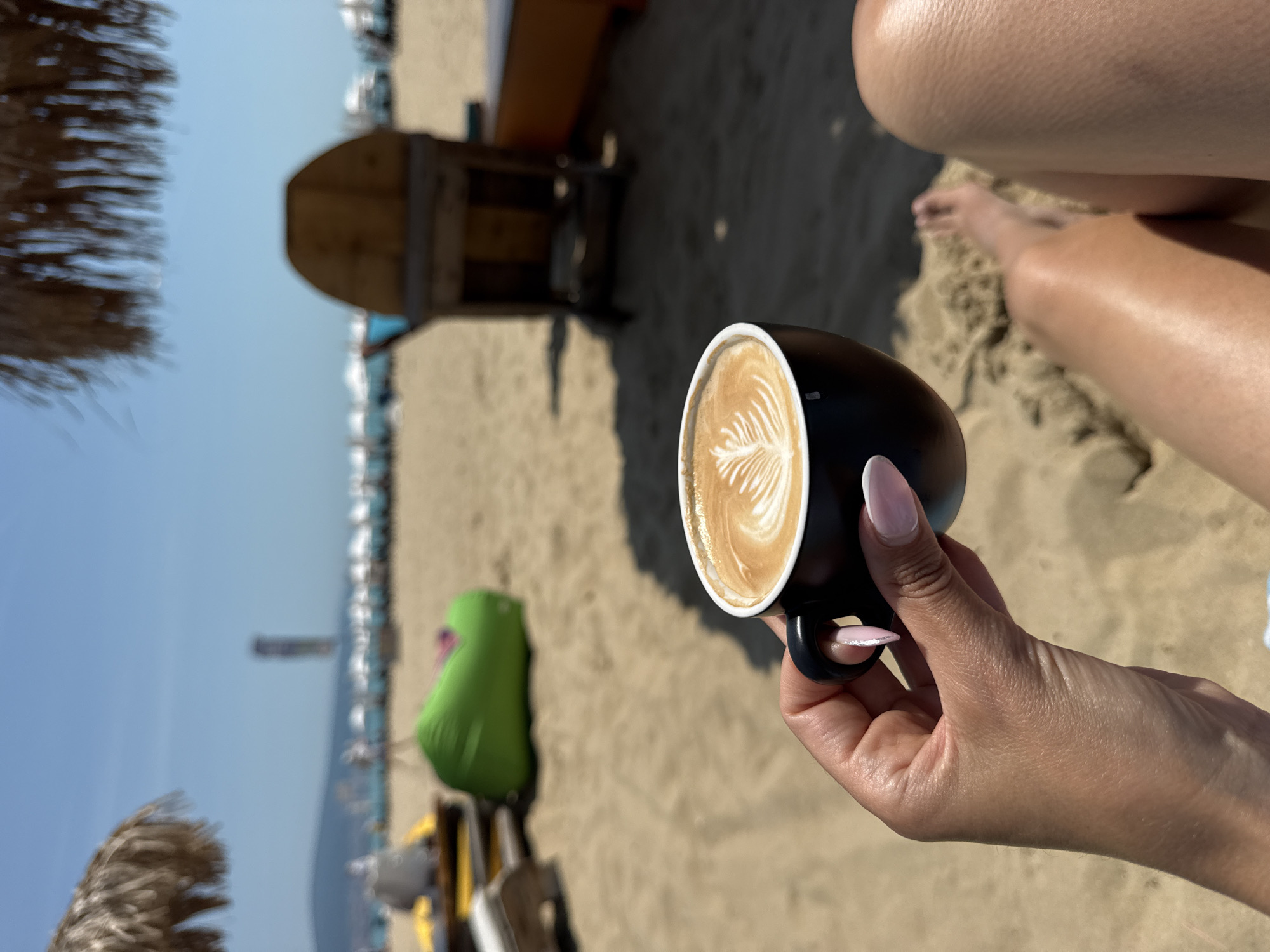 A first-person perspective photograph captures a beach scene in Nesebar, Bulgaria, under bright daylight. In the immediate foreground, a person's right hand, adorned with pink and white ombré manicured nails, firmly grasps a black coffee cup containing a beverage with detailed latte art resembling a fern frond. To the lower right, the person's bare legs, including tanned thighs and knees, are visible, resting on the sandy ground, with a bare foot partially submerged in the sand.
The mid-ground reveals a light brown sandy beach with various beach furnishings. A bright green beanbag chair is positioned to the left of the center. To the far left, a portion of a straw beach umbrella is visible. Further right, a dark wooden beach chair or structure casts a prominent shadow on the sand. In the background, numerous white beach umbrellas and what appear to be lounge chairs are sparsely distributed along the distant shoreline. A tall, dark, possibly inflatable water sports object stands vertically in the far distance, silhouetted against a clear blue sky. A low mountain range forms the horizon line beneath the sky.