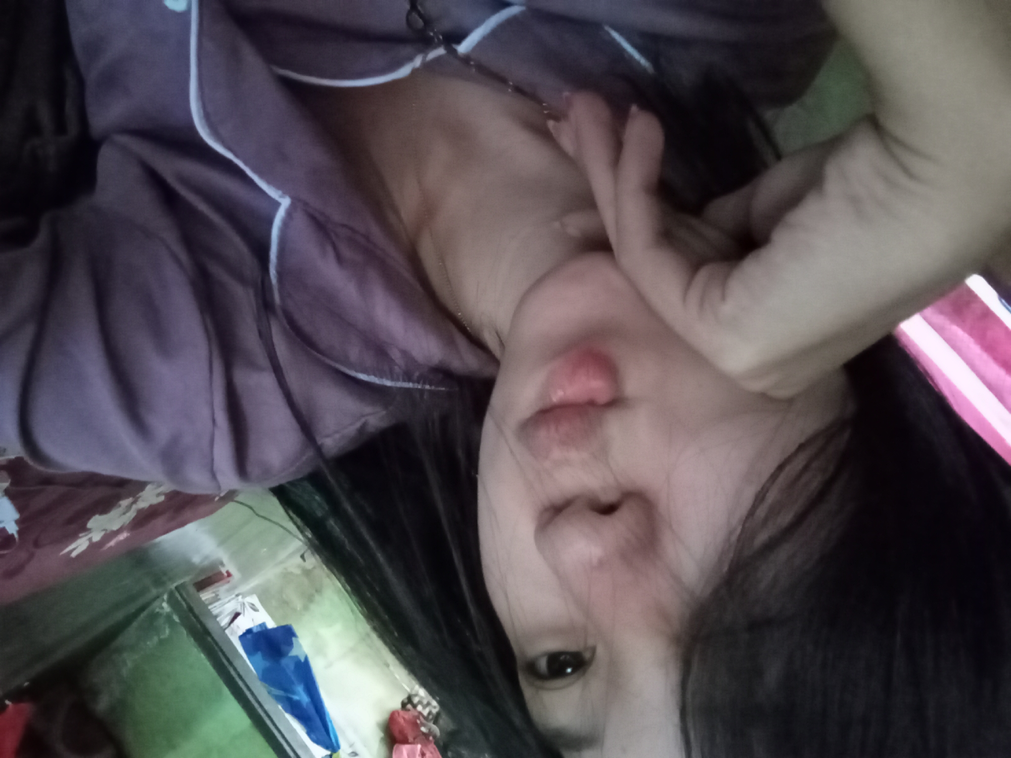 The image shows a close-up, angled shot of a young woman's face and upper body. She has dark hair and is wearing a purple pajama set with white trim. Her right hand is resting on her cheek, and she appears to be looking towards the camera with a soft expression. The background is blurred, but it suggests an indoor setting, possibly a bedroom, with some indistinct objects and colors visible. The lighting is soft and natural. The woman's pose and the overall mood suggest a casual and relaxed moment.