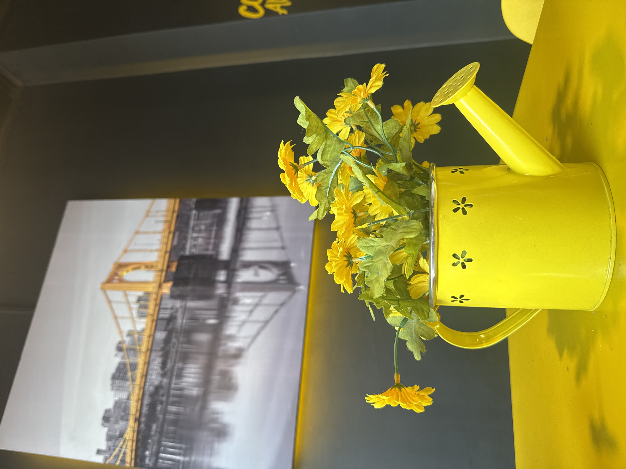 The image, captured from a slightly low angle, displays a bright yellow metal watering can, approximately 8-10 inches in height, holding artificial yellow flowers with green leaves. The watering can, situated on a uniformly yellow table, features three visible daisy-shaped cutouts on its side. In the background, a large framed photograph is mounted on a dark grey wall. The photograph depicts a prominent yellow arch bridge spanning a body of water, with a blurred cityscape reflected in the water; the bridge maintains its yellow color while the surrounding elements are rendered in grayscale. The walls surrounding the scene are dark grey. On the far right wall, partial yellow lettering, possibly spelling "COFFEE" or similar, is faintly visible. The setting is identified as being within the city of Madrid, Colombia. There are no individuals present, and no actions or interactions are depicted.