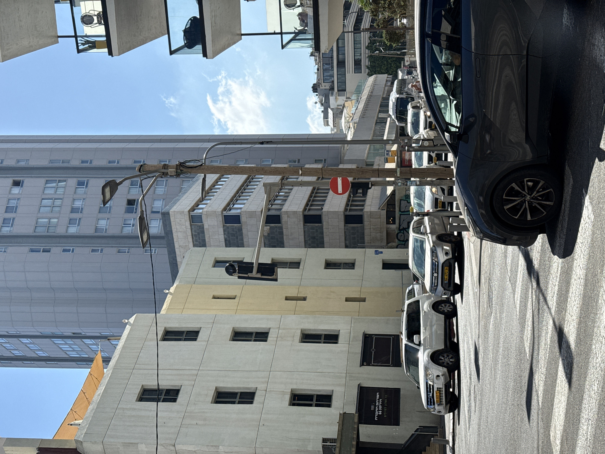The image depicts a street scene in Tel Aviv, Israel, on a clear, sunny day. In the foreground, a dark grey car is partially visible, facing forward, stopped at a crosswalk. The asphalt street shows prominent white crosswalk lines and long shadows cast by streetlights and buildings.
Behind the foreground car, several other vehicles are lined up. A white SUV with a license plate visible is to the left, and a smaller silver SUV is to its right. Further back, other cars and a white van are visible in traffic.
The background is dominated by urban architecture. To the left, a white building with multiple windows and a yellow-colored section stands prominently. Above this, a tall, grey, modern high-rise building with many windows rises into the blue sky. To the right, balconies of a residential building are visible, featuring glass railings and outdoor furniture. Other buildings, varying in height and architectural style, line the street, suggesting a bustling city environment. A traffic light and a street lamp are also visible, along with a circular red and white traffic sign. The overall impression is of a typical urban intersection in Tel Aviv.