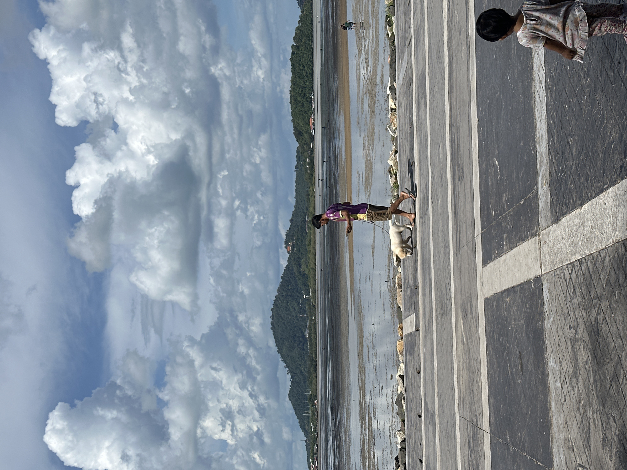 A person is walking a white dog on a paved walkway. The person is wearing a purple shirt with yellow trim and brown shorts, and the dog is on a leash. In the background, there is a body of water with a muddy or sandy bottom, suggesting it may be low tide. Beyond the water, there are green, tree-covered hills and some buildings. The sky is filled with large, white cumulus clouds. In the foreground, to the right, a young child is standing and facing away from the camera, looking towards the water. The walkway appears to be part of a public area, possibly a promenade or park, located near the coast in Phuket City Municipality, Thailand, given the characteristic landscape.