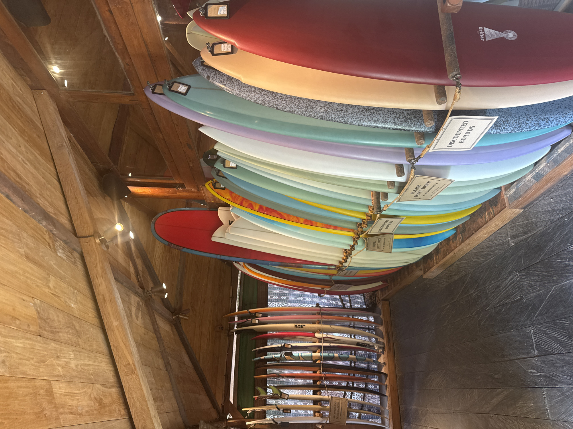 The image shows a display of surfboards in a shop. The surfboards are arranged on racks, with some standing upright and others leaning against the wall. The shop has a rustic wooden ceiling and walls, and the floor is made of dark wood planks. There are several signs visible, including one that says "Please Don't Touch." Another sign reads "Discounted Boards." The surfboards are of various colors and designs, and some have tags attached to them. The overall impression is of a surf shop in a tropical location, possibly Kuta Selatan, Indonesia, given the context.