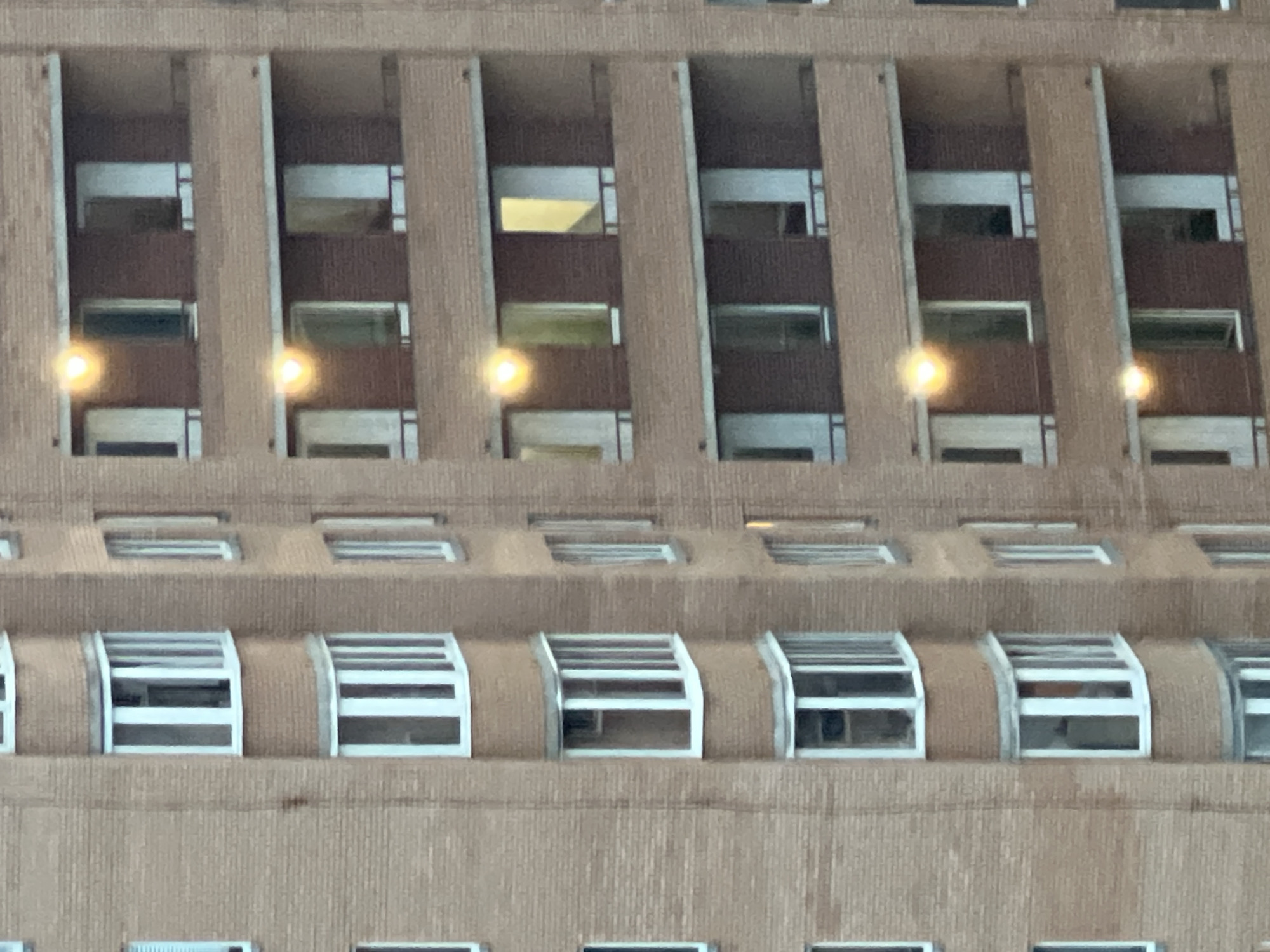 An exterior, eye-level, medium shot captures a brick apartment building with multiple floors. The building is predominantly tan brick with rows of balconies and windows. The balconies are enclosed with white frames and glass, featuring a curved bay window design on some levels. The windows also have white frames, and some are partially open. Lights are visible in some of the windows, creating a warm glow. The building appears to be in an urban setting, as indicated by its scale and construction. The overall lighting suggests it might be late afternoon or early evening.