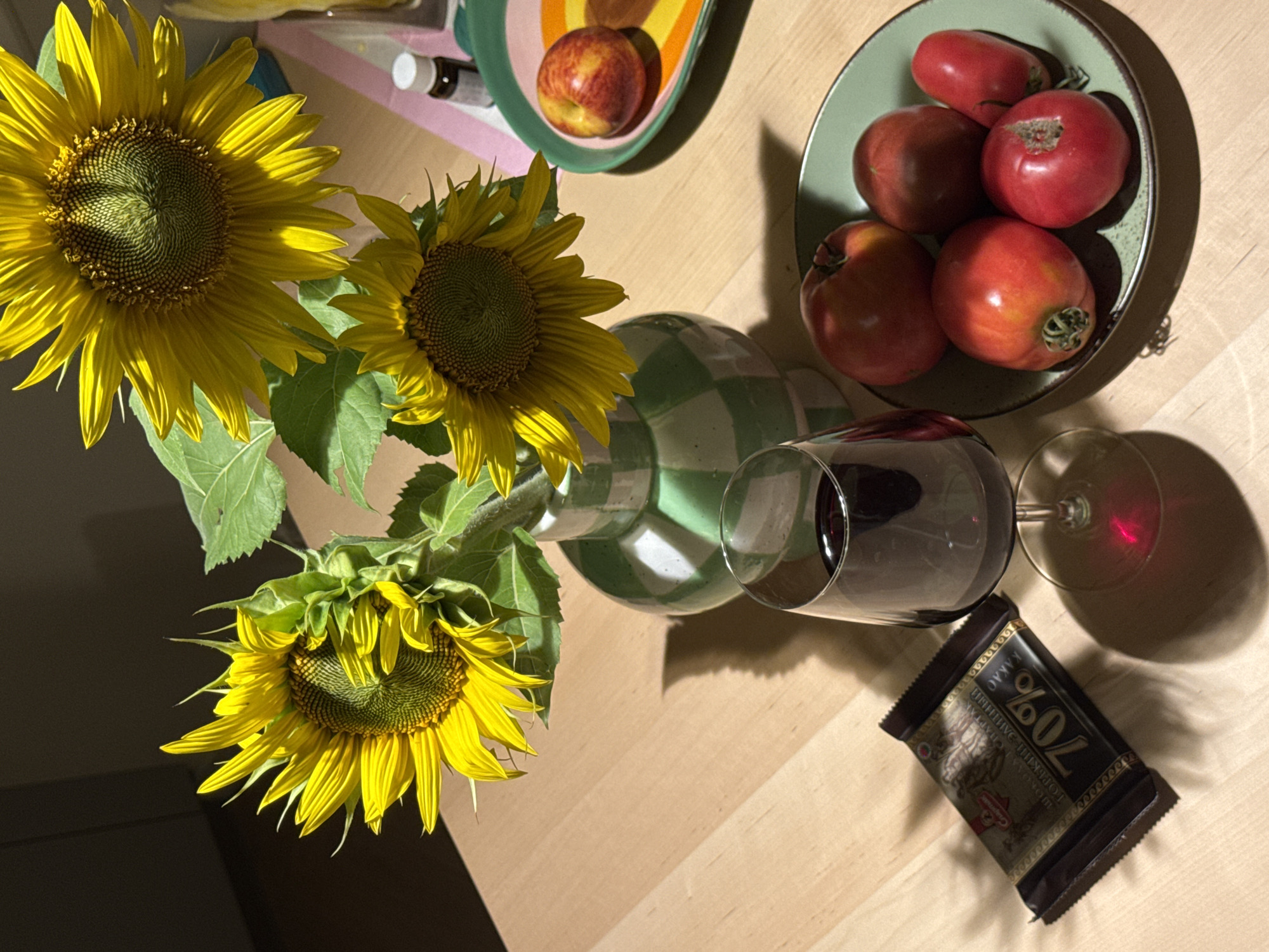 A still life arrangement is depicted on a wooden table, likely indoors. In the center, a green and white checkered vase holds a bouquet of sunflowers. To the left, a dark chocolate bar labeled "70%" lies on the table. Adjacent to the chocolate bar is a wine glass filled with red liquid. To the right of the vase, a small plate holds a red and yellow fruit, possibly a peach or nectarine, and a small dark bottle with a white cap. In the foreground to the right, a plate contains five red tomatoes of varying shapes and sizes. The scene is illuminated by artificial light, casting shadows on the table. The presence of typical household items suggests a domestic setting. There is no indication of the specific location within Izhevsk, Russia.