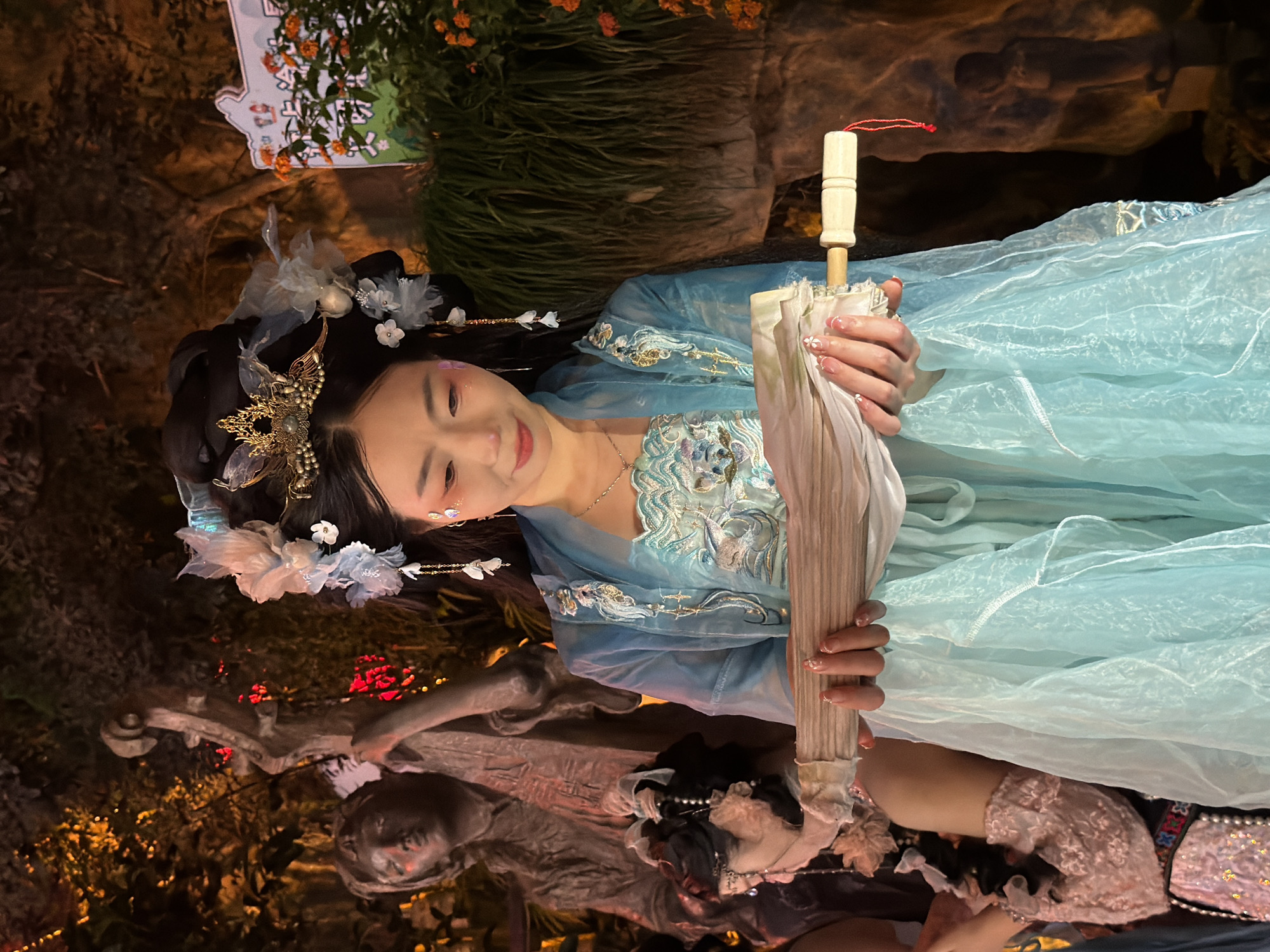 In the city of Yuzhong District, China, a young woman with dark hair adorned with a golden crown and delicate white and blue floral decorations, is the central subject of this photograph. She is wearing a traditional light blue garment with intricate embroidery, and a sheer blue overcoat. Her makeup includes light pink blush and subtle shimmer on her cheeks. She is holding a closed, light-colored umbrella with a light wooden handle in both hands, her fingers interlaced around its shaft. The woman is looking downwards with a gentle expression. In the background, a statue of a person playing a stringed instrument is visible, partially obscured by foliage and warm, reddish lighting. To the right, a lush, green plant with small orange flowers and a sign with Chinese characters are present. The overall setting appears to be an outdoor event or garden, with atmospheric lighting creating a somewhat mystical ambiance.