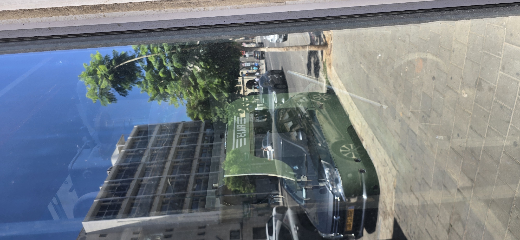 The image shows a reflection in a window of a street scene in Tel Aviv, Israel. In the foreground of the reflection is a dark green car, likely a hatchback, parked on the side of a paved street. The car has its headlights on, and part of its license plate is visible, appearing to be yellow and black. Behind the car, a green truck with the word "ELNA" in white, stylized lettering is reflected. The truck appears to be a commercial vehicle, possibly a delivery truck.
Further in the background of the reflection, a multi-story building with a light-colored facade and numerous windows is visible, characteristic of urban architecture in Tel Aviv. Trees with green foliage are also reflected, suggesting a street lined with vegetation. The sky in the reflection is bright blue, indicating a clear day.
The scene is captured through a window, which introduces reflections of the interior, such as a partial view of the window frame on the right side of the image, which is textured and appears to be made of concrete or stone. There are also some minor smudges and scratches on the glass, common to window surfaces. The street itself appears to be paved with interlocking square stones, a common sidewalk material. Several other vehicles and a few indistinct figures of people can be seen in the reflected scene on the street. The overall impression is a snapshot of a typical sunny day in an urban environment in Tel Aviv.