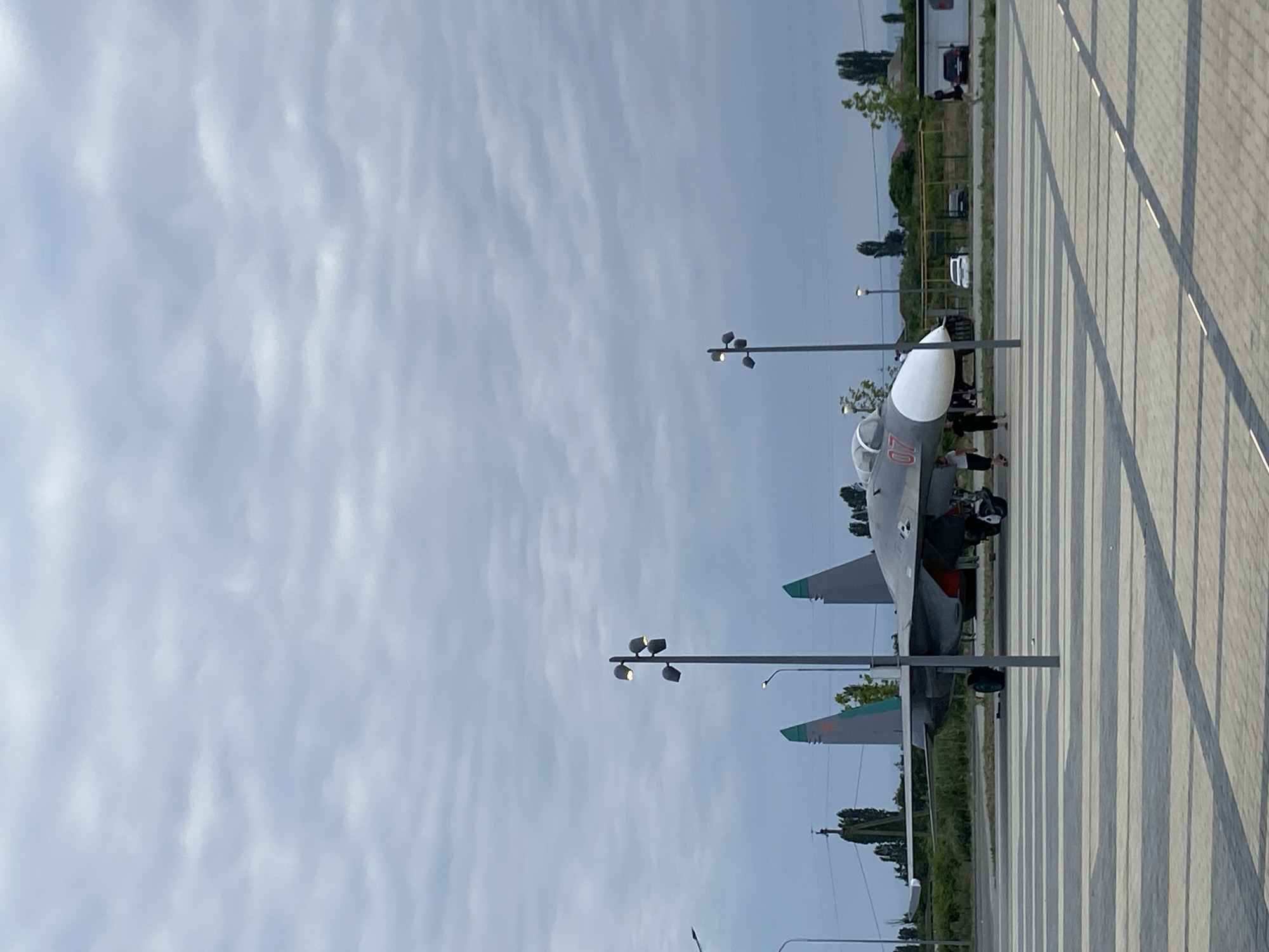 In Arablyar, Russia, a parked military jet with the number "07" visible on its nose cone is displayed on a paved area. The jet is gray with green accents on its tail fins and a white nose cone. Two streetlights are positioned in the foreground, casting shadows on the patterned paving stones. A few people are walking near the jet. In the background, there are trees, buildings, and several parked cars. The sky is overcast with gray clouds.