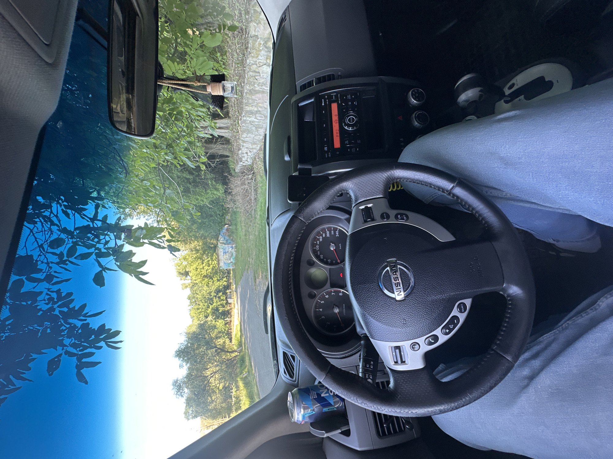 The image is taken from inside a Nissan vehicle, looking out through the windshield. The dashboard is visible, featuring a steering wheel with the Nissan logo, the instrument cluster, and a car stereo. A can of energy drink is placed in a cup holder on the left side of the dashboard. Outside the car, a road leads through a rural or semi-rural area with trees and greenery on either side. There is a stone wall and some graffiti on a small structure in the distance. The sky is clear and blue, with tree branches casting shadows on the windshield. A rear-view mirror hangs from the top of the windshield, with a car air freshener attached to it. The image is a point-of-view shot from the driver's seat. The location is Makiv, Ukraine.
