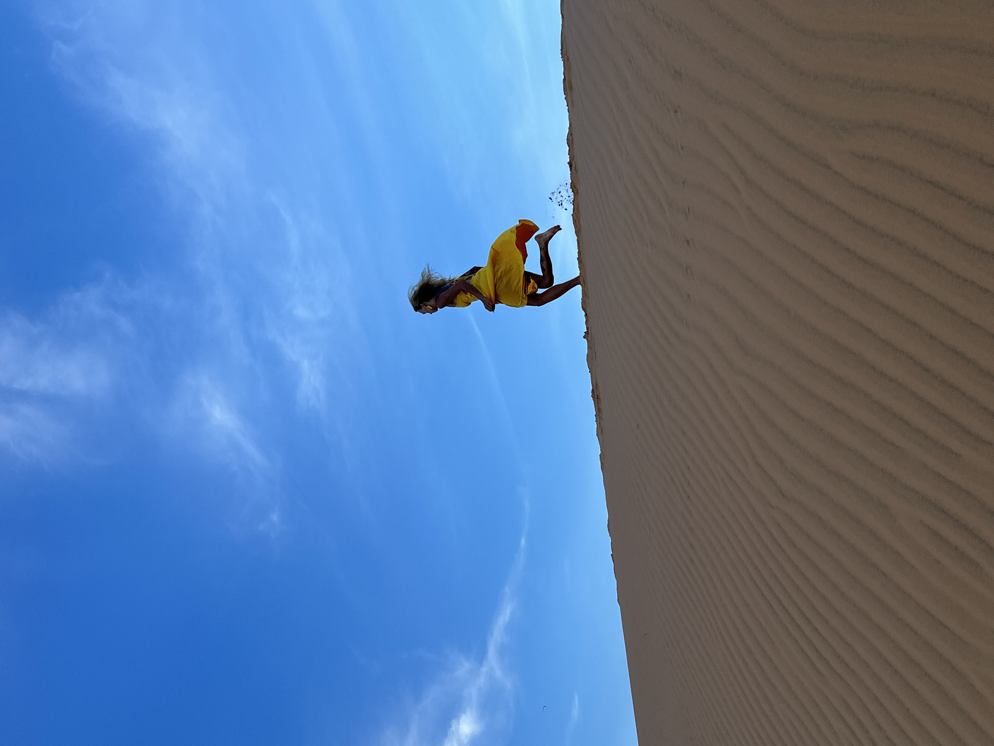 The image shows a person in mid-air, appearing to be jumping or falling down a sand dune. The person is wearing a bright yellow dress, which is flowing behind them. Their hair is also being blown by the wind, suggesting a dynamic movement. The background is a clear blue sky with a few wispy white clouds. To the right of the person, a large sand dune dominates the frame, its surface textured with parallel lines created by the wind. The angle of the shot is from below, looking up towards the sky and the person. This perspective emphasizes the height of the dune and the freedom of the jump. The scene evokes a sense of exhilaration and adventure, possibly capturing a moment of playful abandon in a desert-like environment. The location is identified as Mui Ne, Vietnam, known for its sand dunes and coastal landscapes. The bright sunlight suggests it is daytime, likely with fair weather. There is no visible text in the image.
