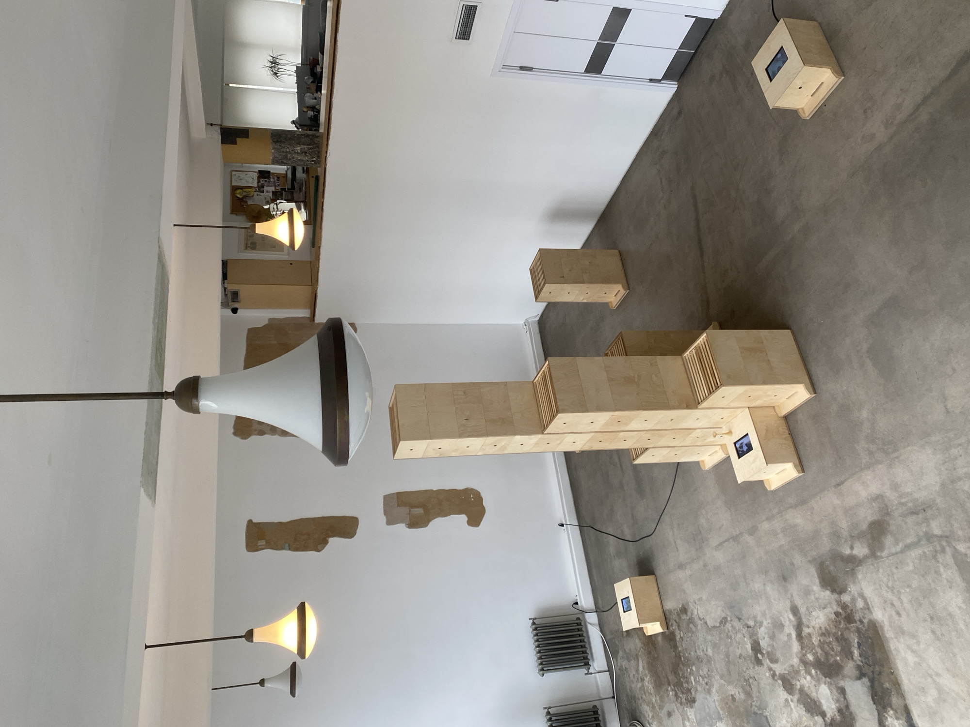 The image depicts the interior of an art gallery or exhibition space, likely in Moscow, Russia. The room has white walls with patches of exposed brick or plaster, suggesting a historical building. Several light fixtures hang from the ceiling, including a large, cone-shaped lamp. The floor is concrete.
A large wooden structure, constructed from stacked wooden boxes, dominates the center of the room. Several smaller wooden boxes with embedded screens are scattered around the floor, possibly acting as interactive displays or monitors. A metal radiator is also visible on the lower portion of the image. There are no people visible in the immediate scene. The overall impression is of a contemporary art installation within a building with architectural character.