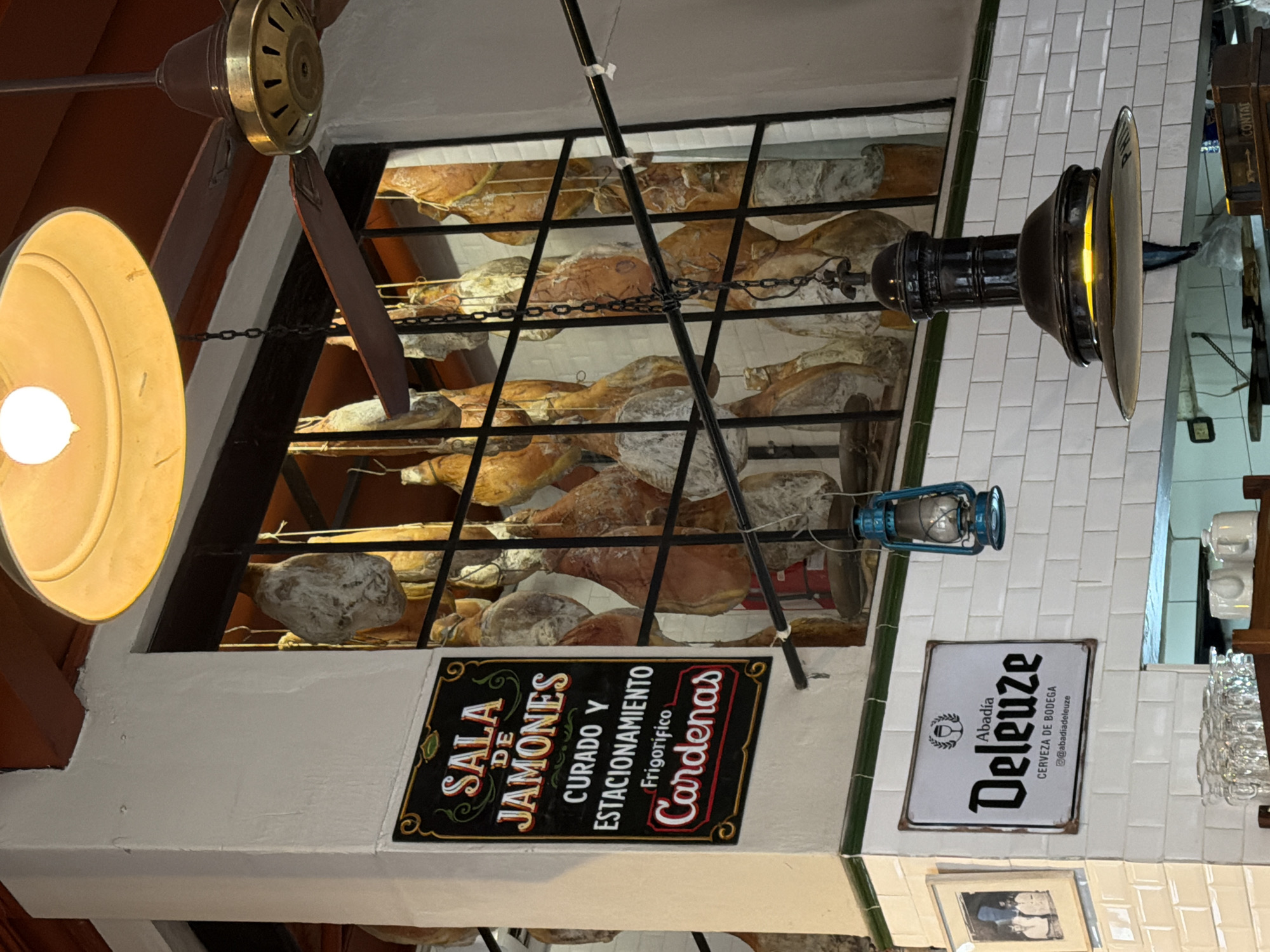 Here is a detailed description of the image:
**Scene:**
The image shows a ceiling view of a business establishment, likely a ham curing and selling location, with additional signage for a brewery. The overall impression suggests a traditional or historical ambiance.
**Objects:**
* **Hams:** Numerous hams are stored on multiple tiers behind a black iron grate.
* **Signage:** Two prominent signs are visible:
* One sign is for "SALA DE JAMONES" (Ham Room) with the text "CURADO Y ESTACIONAMIENTO" (Cured and Stored) and "Frigorífico Cardenas".
* The other sign advertises "Deleuze", a "CERVEZA DE BODEGA" (Craft Beer).
* **Lighting:** Two vintage-style lights are visible:
* A yellow globe-shaped light fixture is present.
* A dark-colored pendant light is positioned closer to the right.
* A small lantern is hanging by a metal rod as decoration.
* **Ceiling Fan:** Part of a ceiling fan is visible in the upper left corner, with brass and brown elements.
* **Wall tiles:** The wall is covered with white rectangular tiles with green trim.
* **Glassware:** A rack of wine glasses.
* **Photograph:** A framed black and white photograph.
**Environment:**
The setting suggests a deli, restaurant, or store specialized in cured hams, with a partnership or advertising for a specific beer brand.
**Location in Buenos Aires, Argentina:**
Based on the presence of "Cardenas" (a possible business name), the local style of signage, and the type of establishment, the location is likely in Buenos Aires, Argentina.