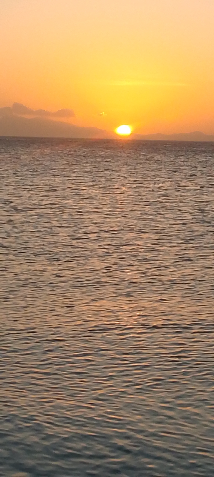 A serene seascape is bathed in the warm glow of a sunset. The sun, a bright orb, hovers just above the horizon line, reflecting on the gently rippling water surface. Distant landmasses, possibly islands or hills, are silhouetted against the bright sky, creating a soft horizon. The sky transitions from a vibrant yellow near the sun to a paler orange hue overhead. The sea is calm, with small waves creating a pattern of light and shadow.