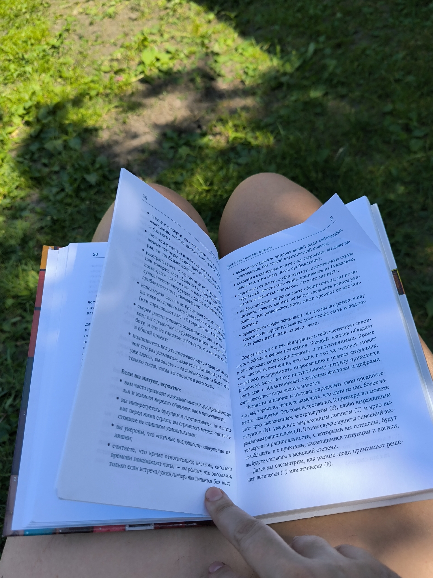 The image shows a close-up of an open book held by a person's legs, which are crossed. The book is open to pages 36 and 37, which contain text in Russian. The person is sitting outdoors, as indicated by the green grass and dappled sunlight visible in the background. The image does not contain any information to determine the location as Maiskii, Russia.