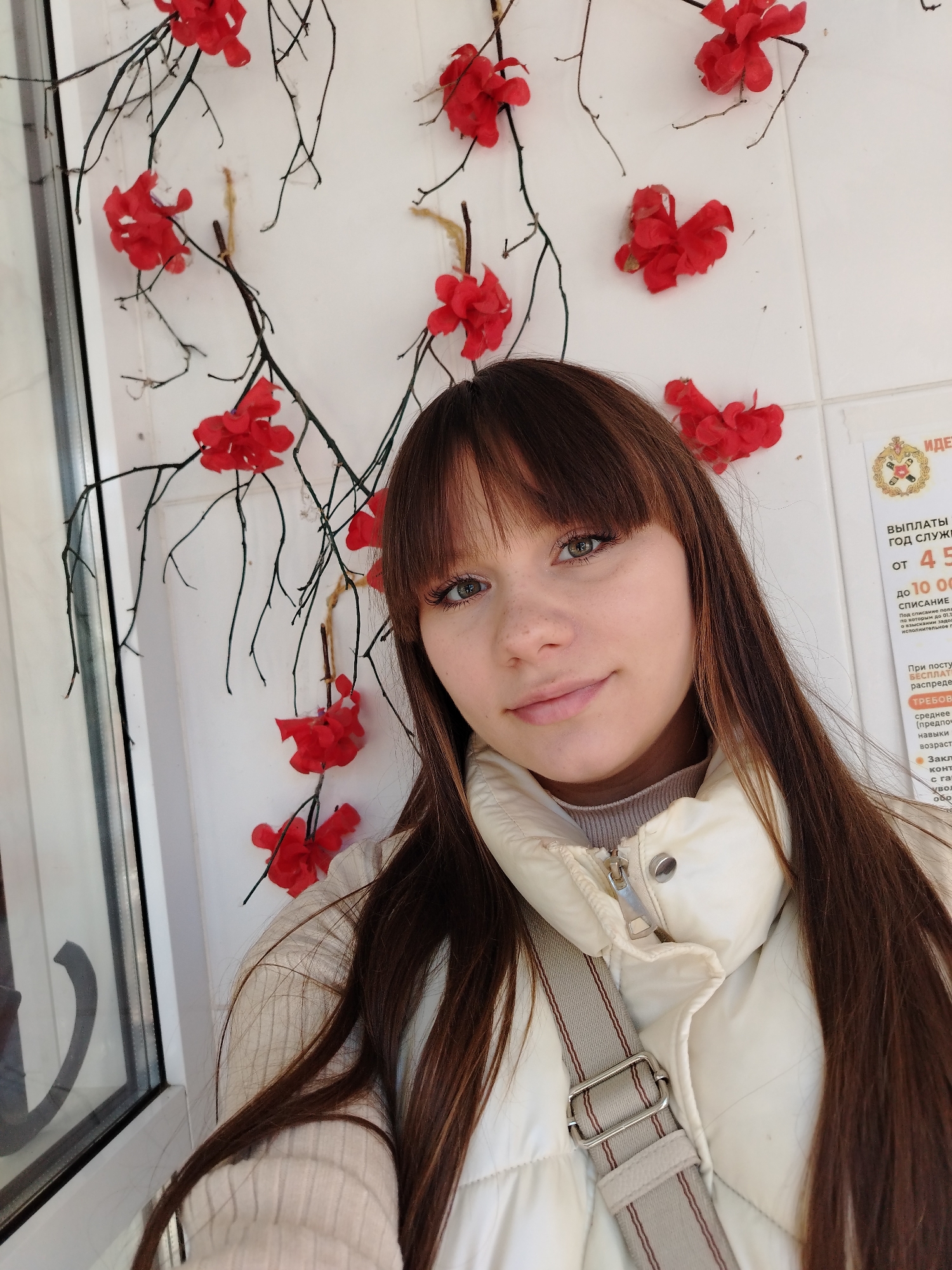 A young woman with long, dark brown hair and bangs is taking a selfie. She has fair skin and is wearing a light-colored padded jacket over a beige sweater. A gray bag strap is visible across her body. She is indoors, standing in front of a white wall decorated with bare branches and bright red, artificial flowers. To the right of the woman, a white sign is visible with text in Russian. The text includes figures "4 5" and "10 00". From the text, the location can be identified as an office in Chaikovsky, Russia, likely related to government services or financial matters.