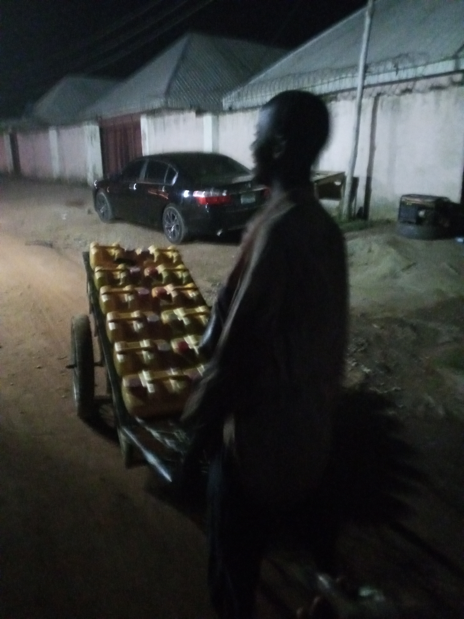 The image appears to be taken at night in Galadimawa, Nigeria. The location is a street with a black sedan parked against a white wall with a gated entrance to a residential property. A man stands next to a two-wheeled cart loaded with multiple jerrycans. The man is in the foreground, partially obscured by darkness. The area is dimly lit.