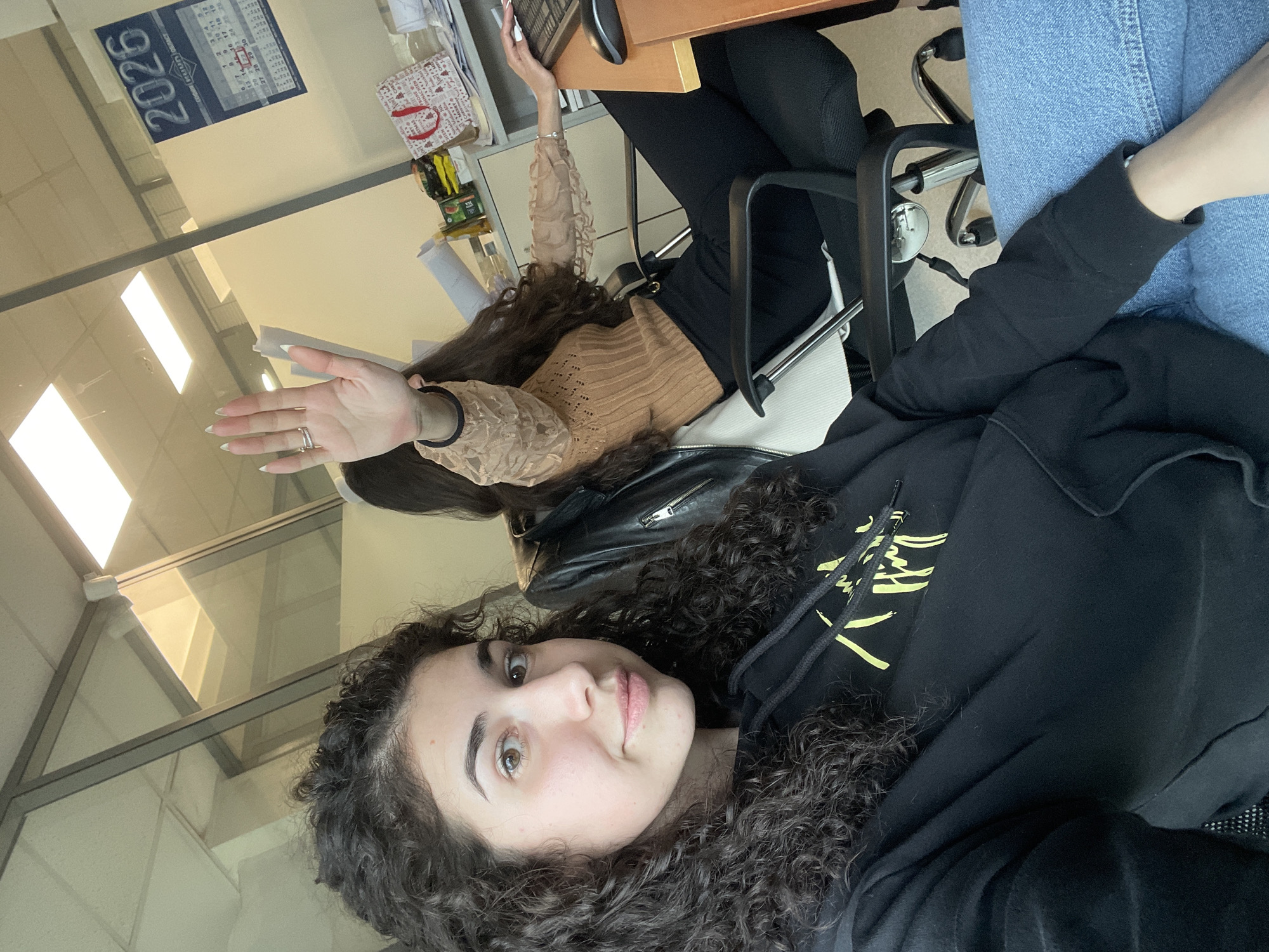 This image, taken in an office setting in Sofia, Bulgaria, depicts two young women. In the foreground, a woman with dark, curly hair looks directly at the camera with a neutral to slight smile. She wears a black hoodie adorned with yellow graphics and blue denim jeans.
Behind her, another woman with long, dark, wavy hair is partially visible. She is seated at a wooden desk, dressed in a light brown, textured long-sleeved top and black pants. Her right hand is raised, displaying long white fingernails and a ring on her ring finger, while her left hand rests on a computer keyboard, suggesting she is working or has paused.
The office environment includes black office chairs with chrome accents, a wooden desk, and glass partitions reflecting bright overhead lighting, indicating it is daytime. On the desk, various office supplies, papers, and a red and white patterned bag are visible. A black leather jacket is also seen draped near the foreground subject.
A wall calendar in the background prominently features the year "2026" and Cyrillic text, including "РЕКА ОБЛА". The scene captures a casual moment in a modern workspace.