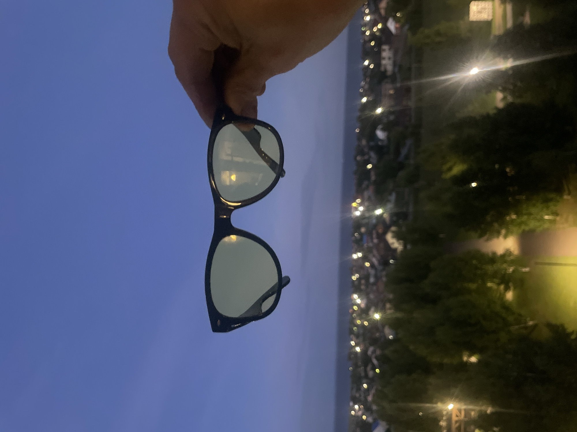 A pair of black-rimmed sunglasses with light green lenses is held up in the foreground by a person's hand against a twilight sky. The background features a blurred, bokeh-effect view of a city at night, with numerous lights from houses and streetlamps visible. Trees are also visible in the mid-ground, some illuminated by artificial light. The general setting appears to be an elevated viewpoint overlooking a residential area.