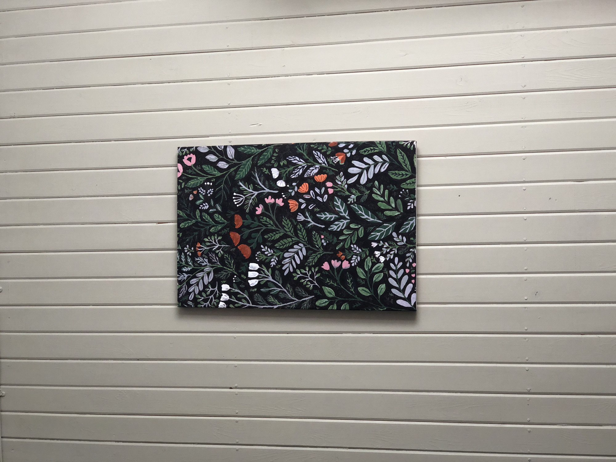 A vertically oriented rectangular canvas featuring a stylized botanical pattern is centrally affixed to a light greige, vertically paneled wall. The painting's background is dark, appearing black, adorned with a dense arrangement of various foliage and floral elements. Leaves are rendered in shades of dark green, medium green, light green, white, and pale lavender-grey. Among the foliage are groupings of white bell-shaped flowers, small clusters of pinkish-purple flowers, and larger, open blossoms in a rust-orange hue. White, star-like structures resembling seed heads are also interspersed. The wall behind the artwork consists of uniformly sized vertical wooden planks with distinct grooves between them. Rows of small, circular indentations, consistent with nail or screw heads, are visible horizontally across the panels. The wall surface exhibits a matte finish with subtle wood grain visible on individual planks. The scene is illuminated evenly, casting minimal shadows. The location is Unknown, Unknown.
