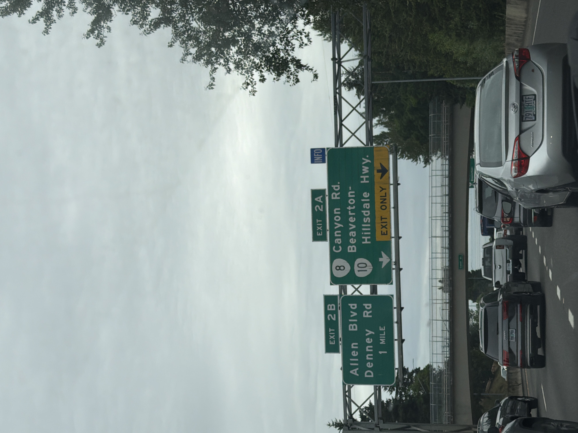 The image shows a view from a vehicle on a highway, likely in Beaverton, Oregon, United States of America, based on the signs. Overhead green highway signs indicate exits and destinations. The left sign shows "EXIT 2B" and "Allen Blvd Denney Rd 1 MILE". The right sign shows "EXIT 2A", with "8 Canyon Rd.", "10 Beaverton-Hillsdale Hwy.", and "EXIT ONLY" with a downward arrow. A blue "INFO" sign is also visible above the right sign.
The scene depicts a multi-lane highway with several vehicles in traffic, including a dark grey SUV in the foreground, a white pickup truck, and a silver sedan. The weather appears overcast. Trees and vegetation are visible along the side of the highway and on an overpass in the background. The overall impression is a typical highway scene during daylight with moderate traffic.