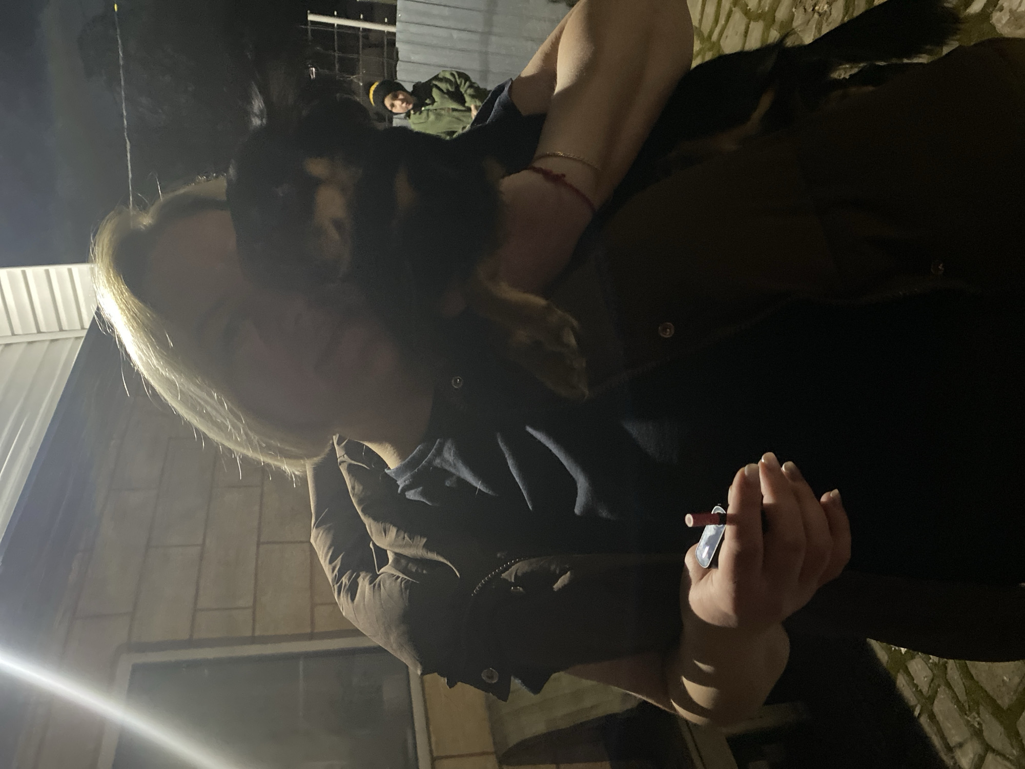 A woman with blond hair holds a small dog on her shoulders. She wears a dark brown jacket. In her hand, she holds a small object, possibly a toy or device. Behind her, partially visible, is another person in a green jacket and a dark hat. The background shows a building with a brick facade and a metal fence. The scene appears to be outdoors at night. The location is Noviy, Russia.