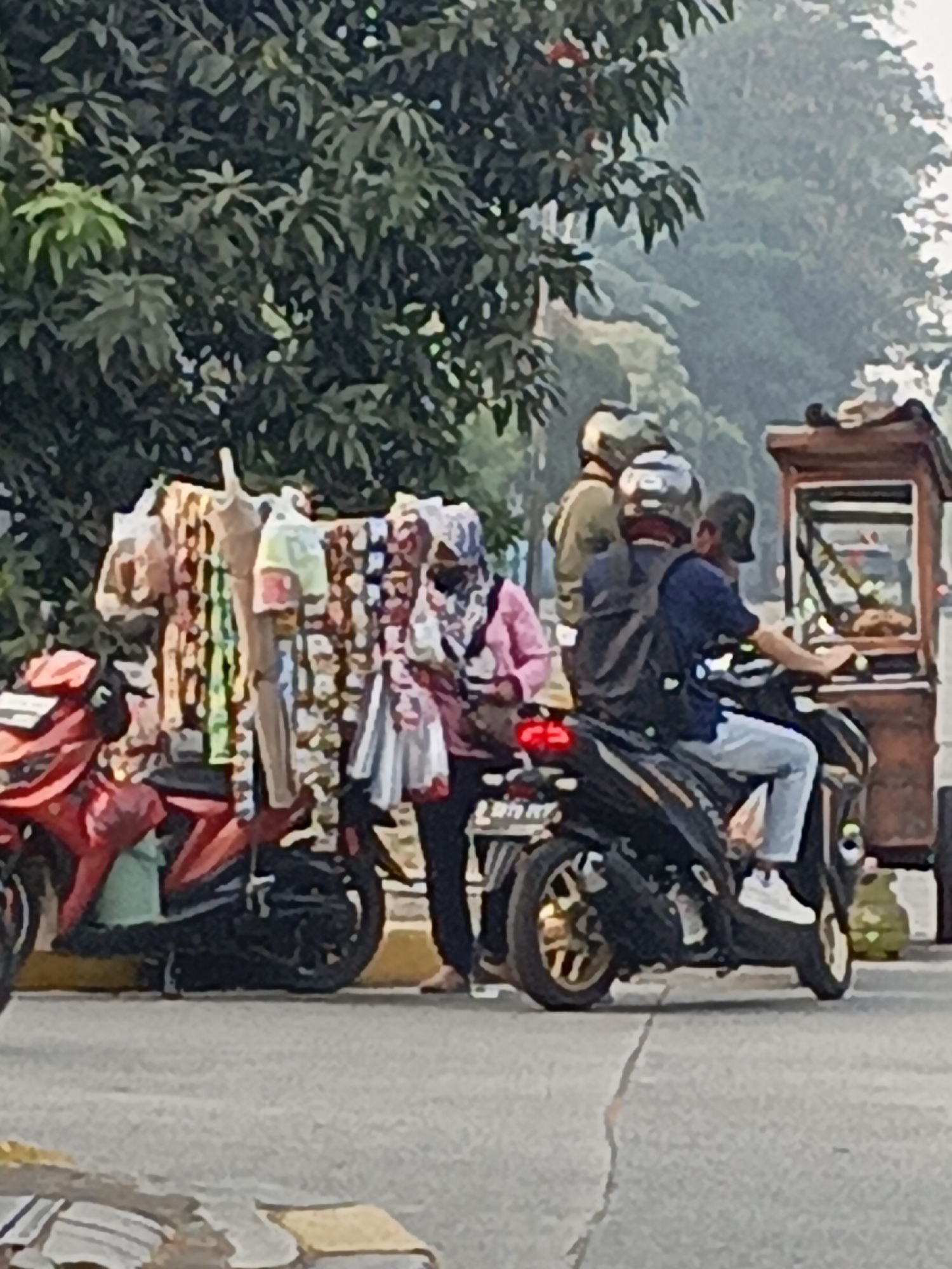 This image, captured in East Jakarta, Indonesia, depicts a street scene with several individuals and vehicles near a food cart. In the foreground, two motorcycles are parked. A red motorcycle is on the left, and a black motorcycle is to its right, with a person wearing a blue shirt and jeans riding it. Behind the motorcycles, a rack displays various clothing items, some appearing to be dresses with colorful patterns. A woman in a pink shirt and a patterned headscarf stands next to the rack. To the right, a food cart with a wooden frame and glass display is visible, with another person behind it, possibly the vendor. In the background, trees and foliage line the street, and a hazy atmosphere suggests either early morning mist or pollution. The overall scene captures a moment of everyday life in a bustling urban environment.