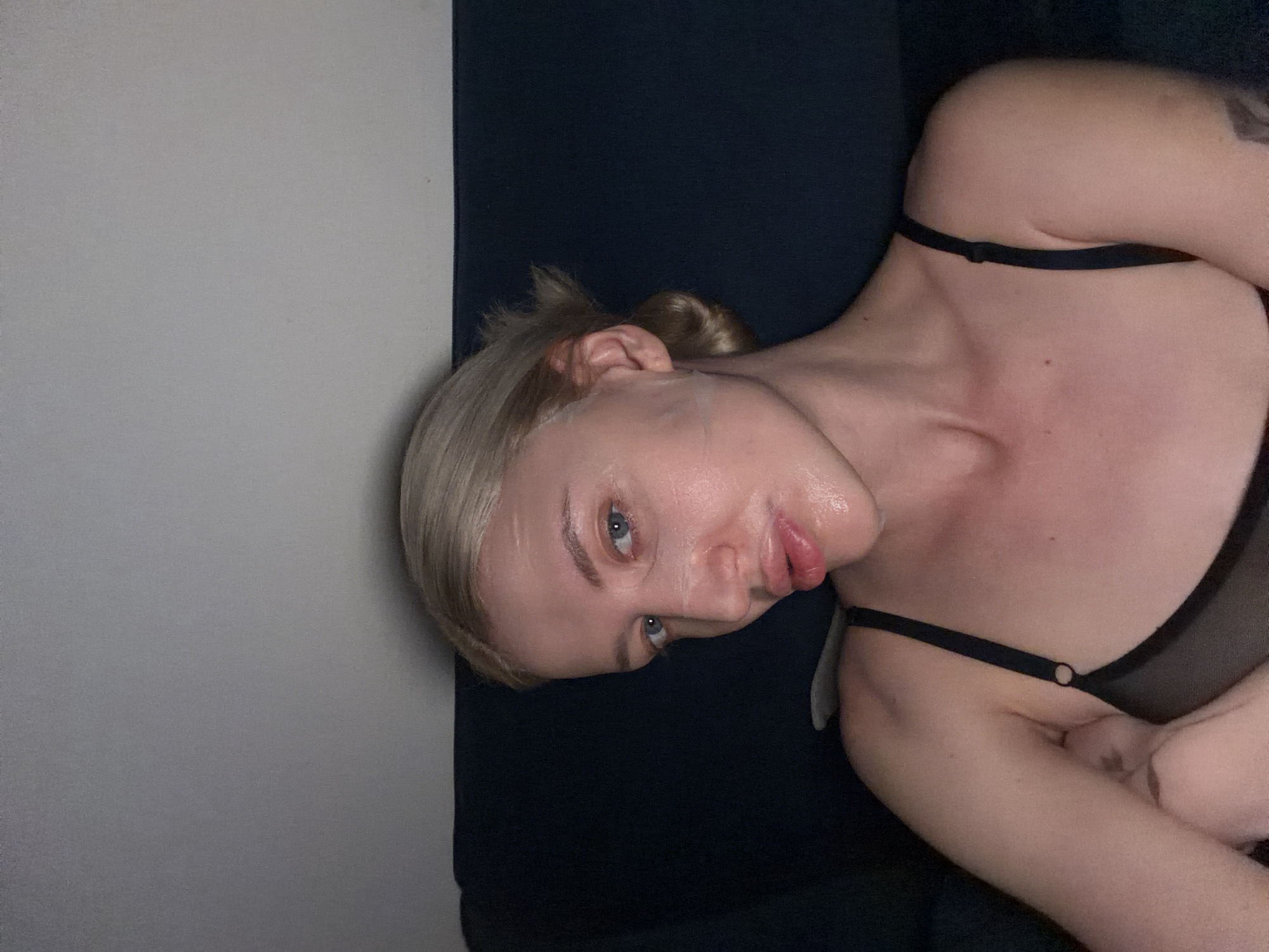 The image shows a blonde woman with blue eyes, fair skin, and a small tattoo on her right shoulder lying on a dark blue or black surface, possibly a couch. Her hair is pulled back in a bun. She is wearing a black bra. The background consists of a light grey wall. The location is impossible to determine from the image.
