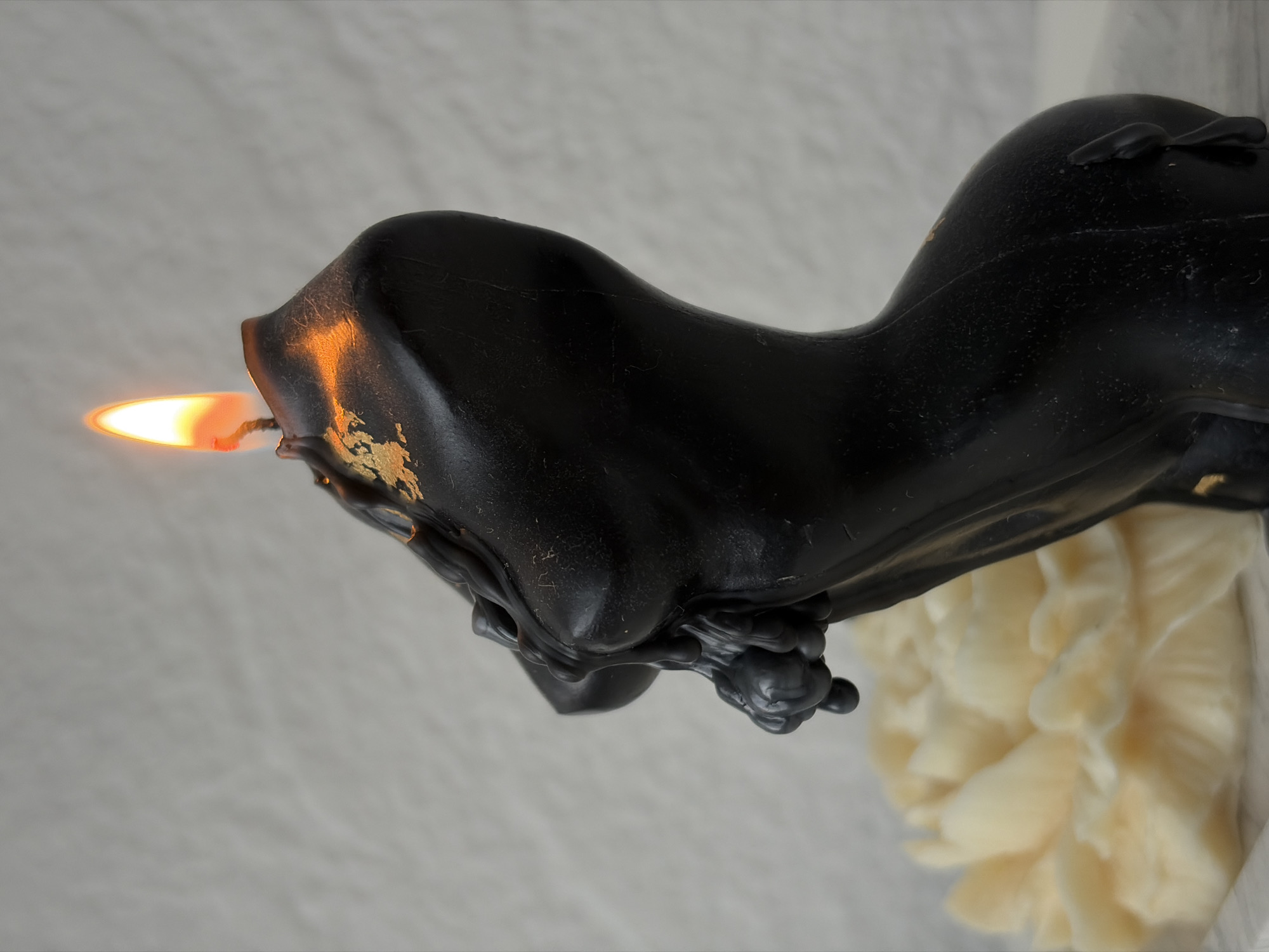 The image shows a lit black candle shaped like a horse head, placed on a white surface with a cream-colored flower-like object underneath. The candle is actively burning, with a prominent orange flame visible. There is no indication of the location being the city of Chaikovsky, Russia, from the image alone.
