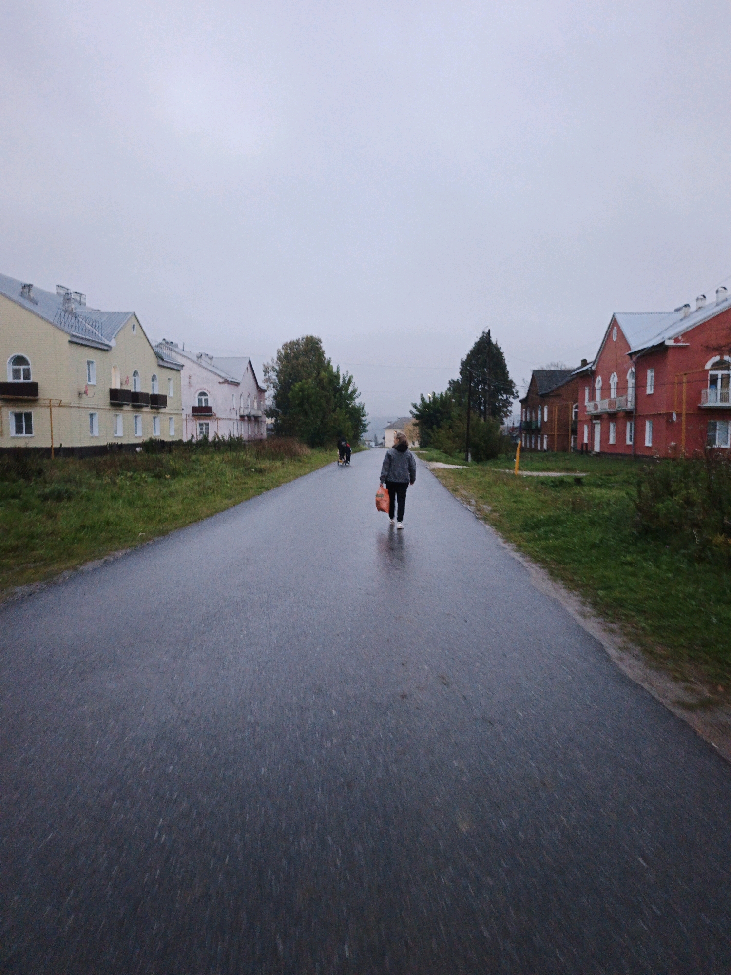 A person walks down a wet, asphalt road in the city of Medvedyovka, Russia. The person, wearing a grey hooded jacket and dark pants, carries a bright orange bag in their right hand. They are walking away from the camera, towards the horizon. To the left of the road are several multi-story buildings with light-colored facades and peaked roofs, some with balconies and windows. Green grass and some trees line the side of the road. To the right of the road, there are red and brown buildings, also multi-story with peaked roofs and windows. A few trees are visible behind these buildings. The sky is overcast and grey. The road appears wet, indicating it may have rained recently or is currently raining lightly. There is a slight blur effect, suggesting motion.