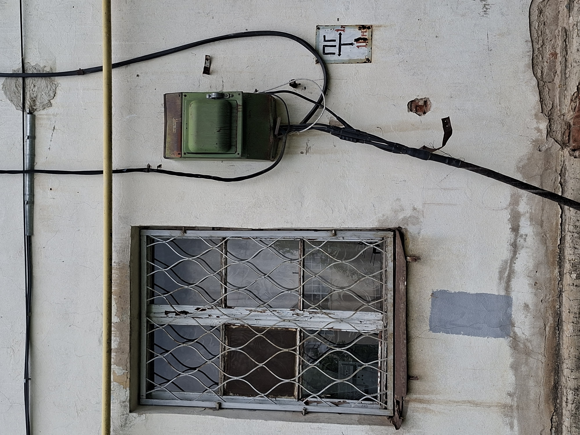 The image shows the exterior of a building in Izhevsk, Russia. The building has a light-colored stucco wall, which appears weathered and shows signs of disrepair, with peeling paint and water stains. A window with a metal grate is visible on the left side of the frame. The grate is made of interwoven curved bars and shows signs of rust. The window itself appears to have multiple panes, some of which are dark or reflective.
Above the window, a yellow gas pipe runs horizontally across the wall. Two black electrical conduits are also attached to the wall, one running horizontally near the top and another running vertically.
On the right side of the wall, a green metal electrical box is mounted. It is weathered and has a hinged lid. Several black electrical cables enter and exit the box, with some appearing bundled and covered in black tape. A smaller white cable is also connected. Below the electrical box, a small rectangular sign with Cyrillic lettering "ПР" and a directional arrow is mounted.
Further down and to the right, a larger black electrical conduit runs diagonally downwards. The wall shows further signs of damage, including a small hole revealing brickwork and areas of crumbling stucco, particularly near the bottom of the image. A patch of grey paint is visible on the wall below the window. The overall impression is one of an older, utilitarian structure in an urban environment.