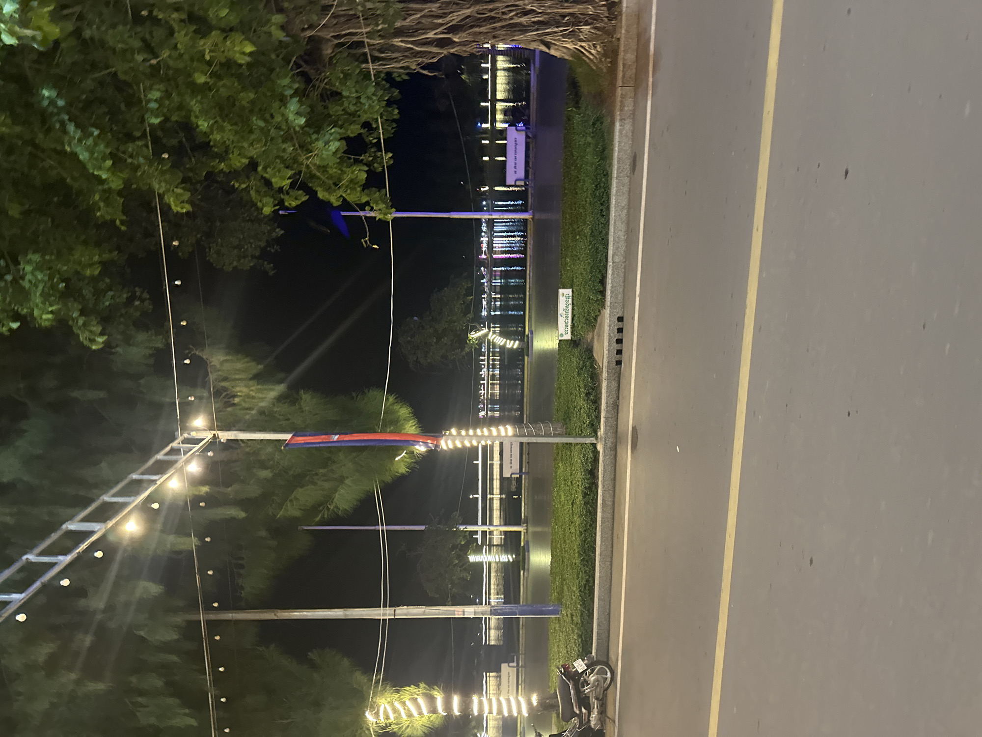 This image captures a nighttime scene along the Kampot River in Kampot, Cambodia. A street runs across the foreground, marked by a yellow line and a white line separating it from a planted area. A parked motorcycle is visible on the left side of the image, near the planted area. Beyond the planted area lies the Kampot River, its surface reflecting the lights from structures and decorations along the riverbank. Trees line the river, and some of them are illuminated with decorative lights. A flag with red and blue stripes is prominently displayed on a pole. The scene is illuminated by streetlights and decorative string lights, creating a festive atmosphere.