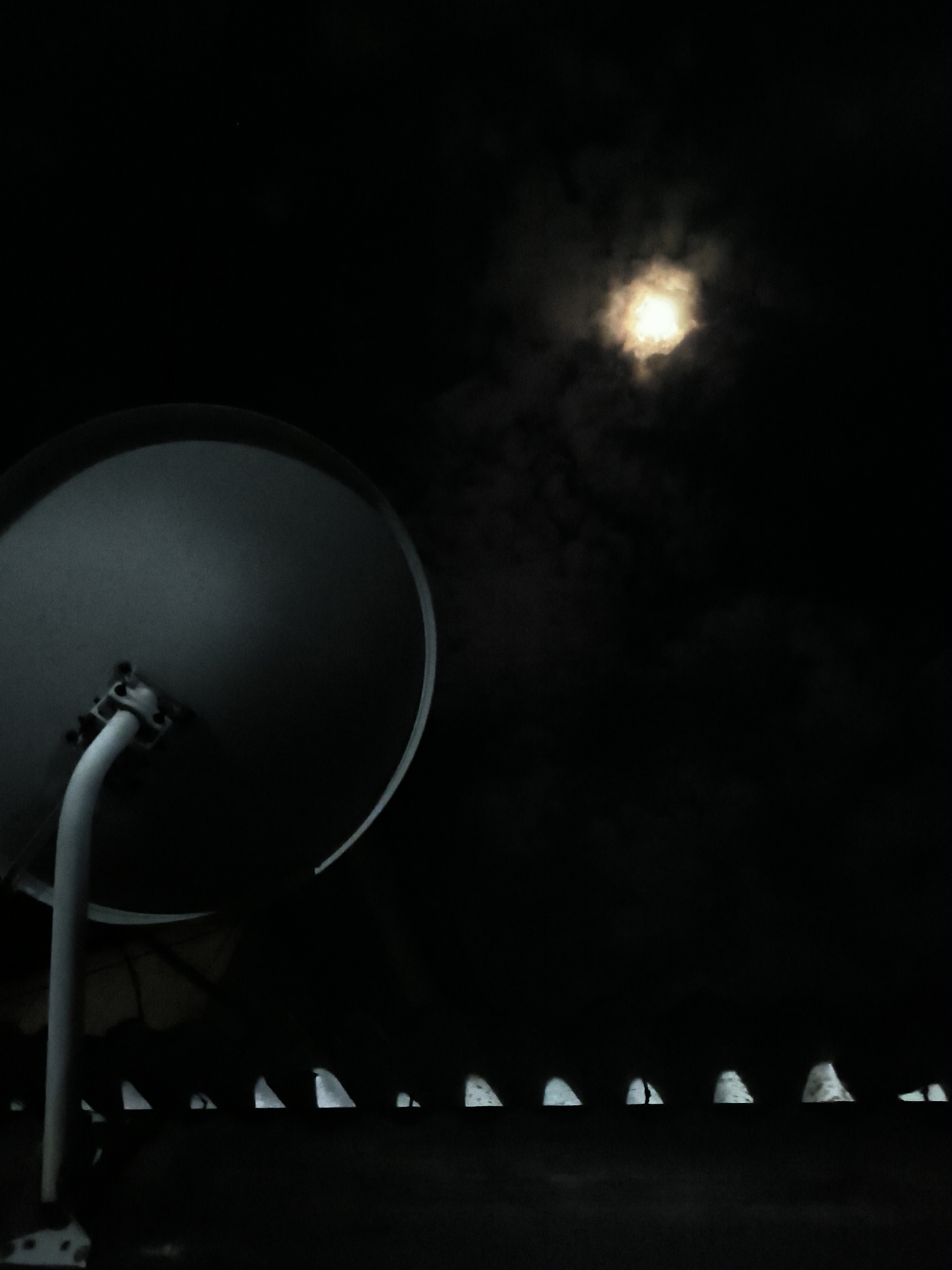 A nighttime scene features a satellite dish in the left foreground with its supporting arm and base visible. Behind the dish, a row of dimly lit, white triangular shapes lines the horizon, possibly the tops of buildings. Above them, the moon, partially obscured by clouds, provides the only significant source of light, casting a diffused glow. The location is Lhokseumawe, Indonesia.